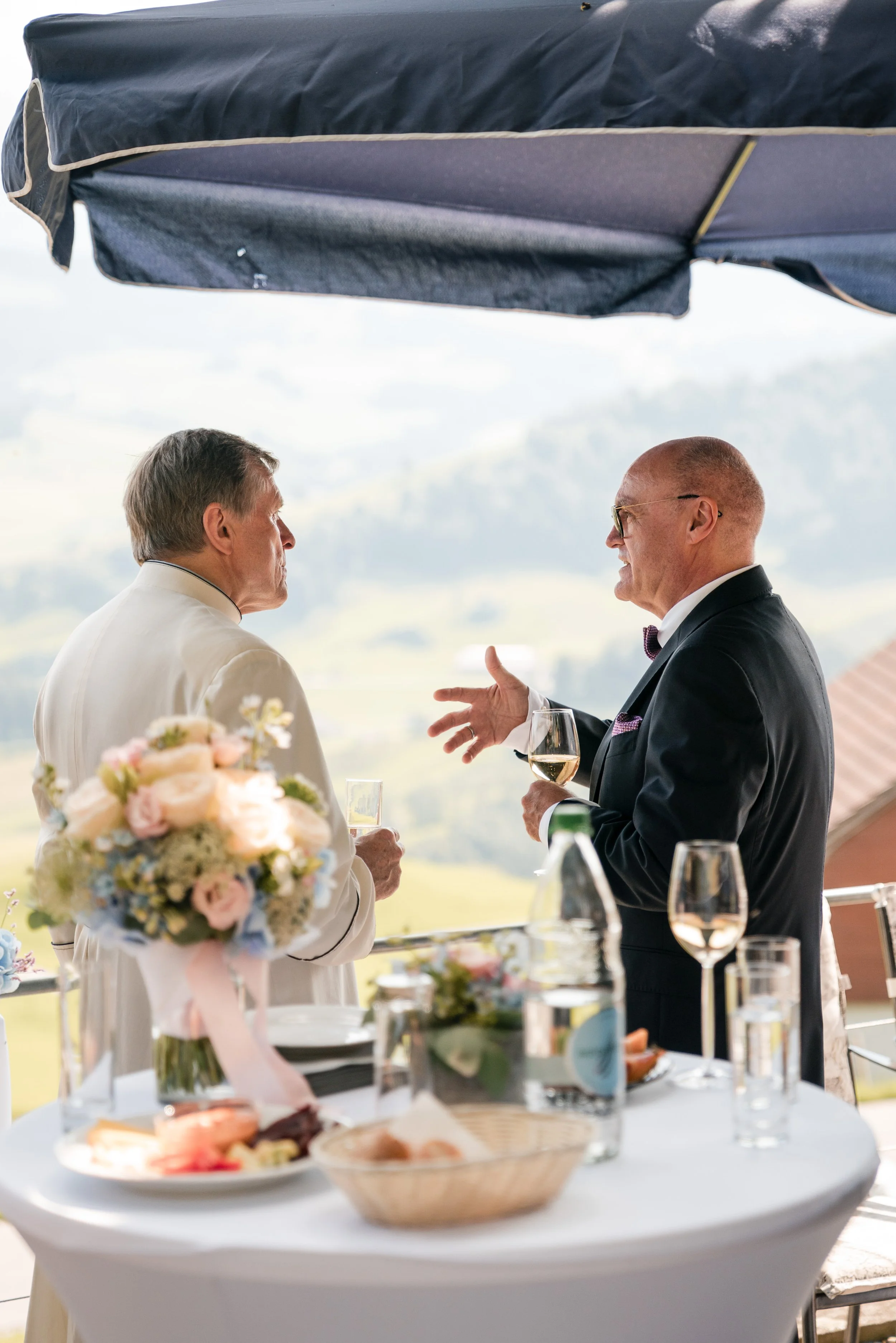 Zwei Männer bei einem Gespräch auf einer Terrasse bei einem festlichen Anlass, mit einem Tisch voller Blumen, Speisen und Getränke im Vordergrund, im Hintergrund eine ländliche Landschaft.