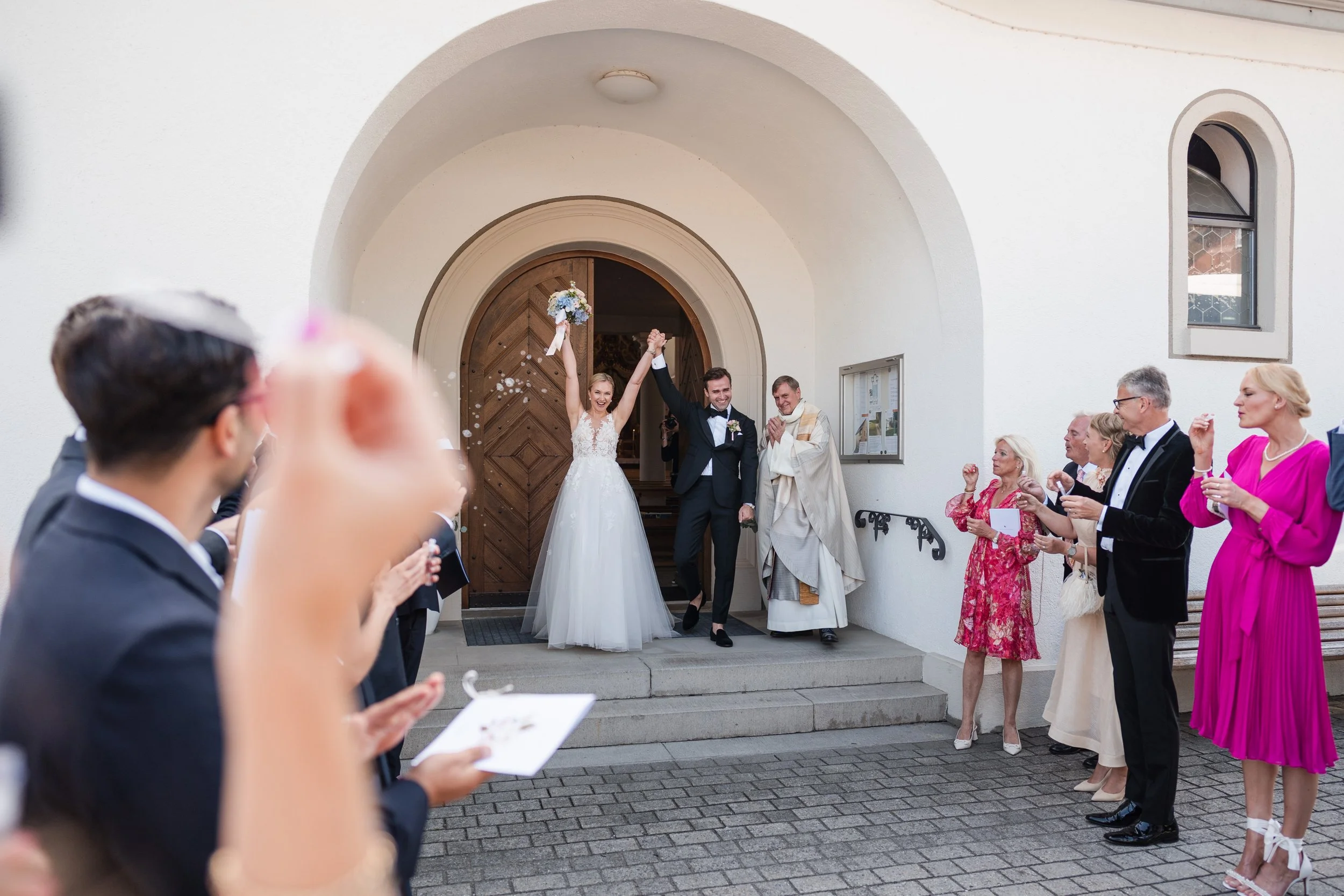 Braut und Bräutigam feiern ihren Hochzeitstag vor der Kirche, umgeben von Freunden und Familie, während sie aus der Kirche kommen. Die Braut trägt ein weißes Hochzeitskleid, der Bräutigam einen dunklen Anzug. Gäste werfen Konfetti.