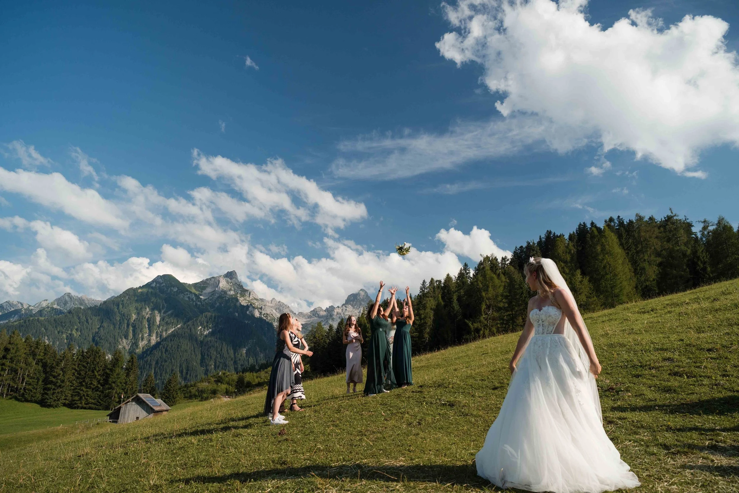 Eine Frau in Brautkleid und mehrere Frauen in Abendkleidern, die auf einer grasbewachsenen Wiese im Freien unter einem blauen Himmel mit Wolken stehen, während sie einen Brautstrauß werfen.