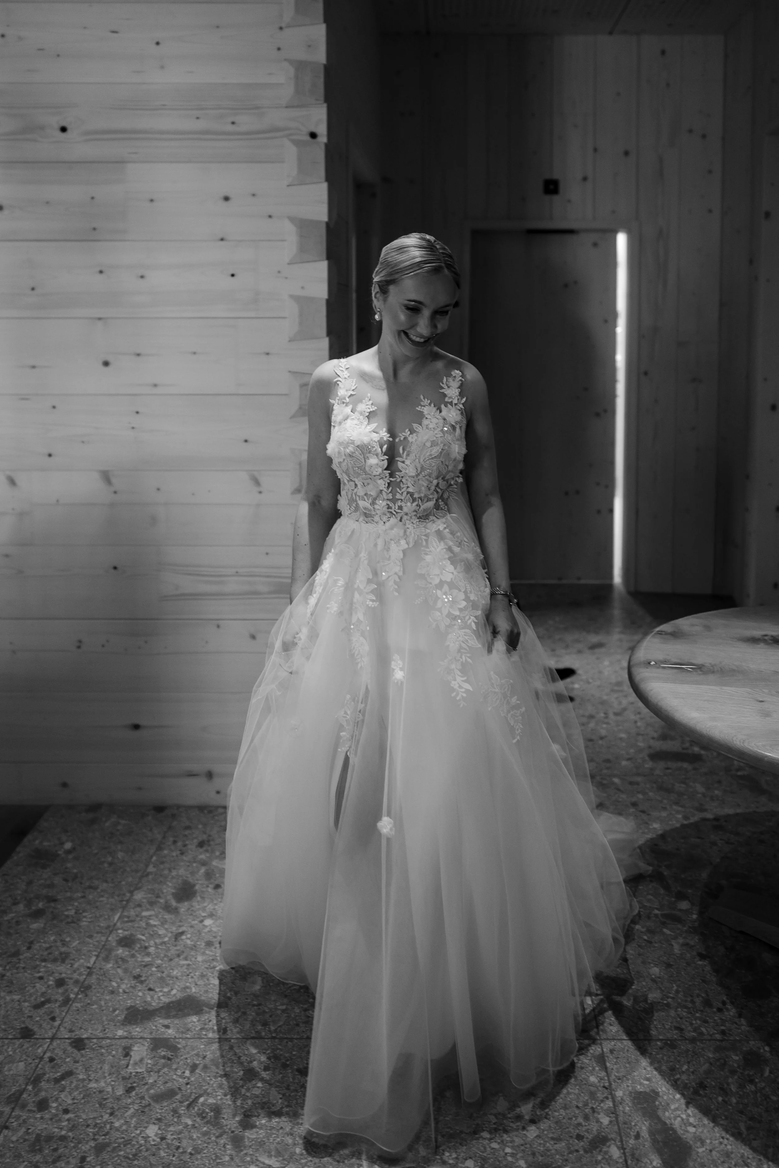 Frau in Hochzeitskleid lächelnd, steht in einem Raum mit Holz-Wänden, schwarz-weiß Foto.