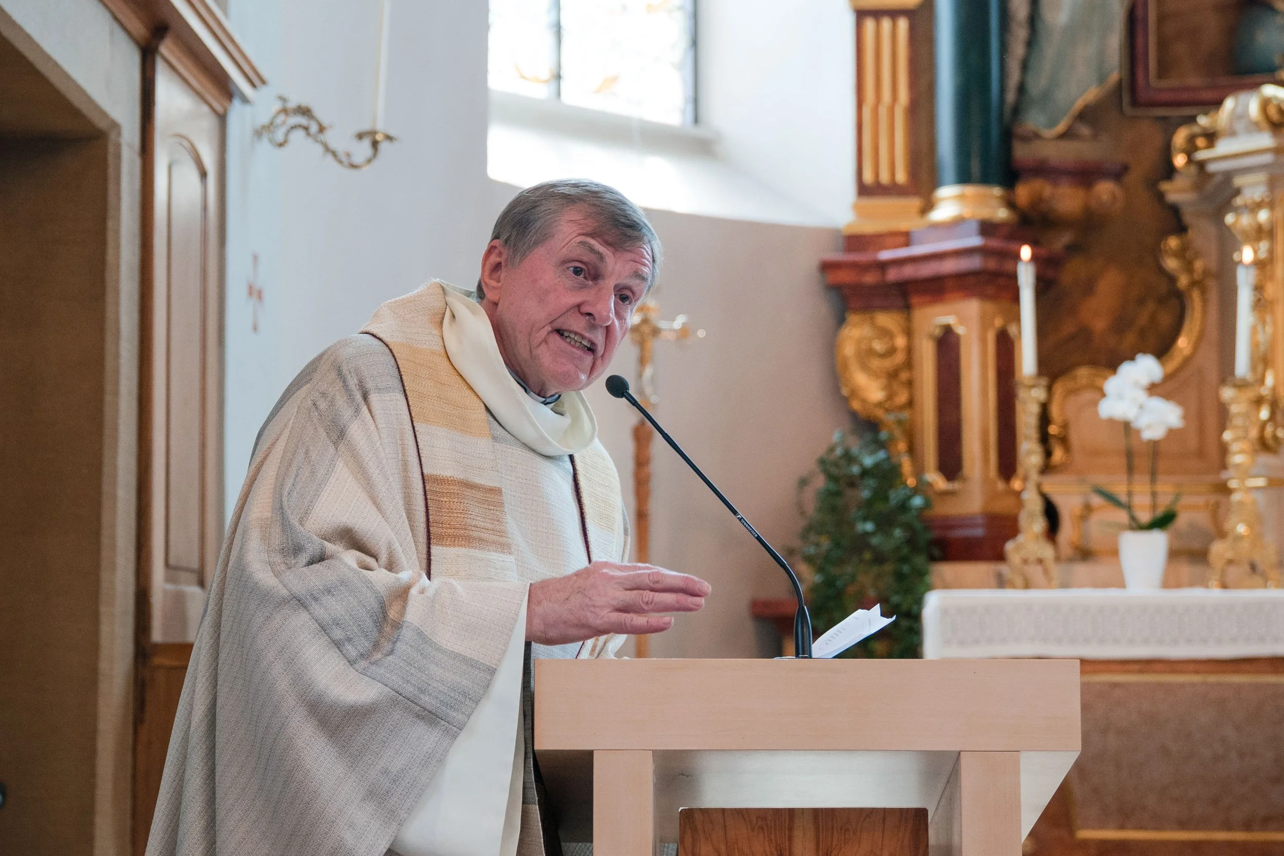 Ein Priester spricht während einer Messe in einer Kirche, stehend hinter einem Holzpodest mit Mikrofon, mit goldverzierten Altargerät im Hintergrund.