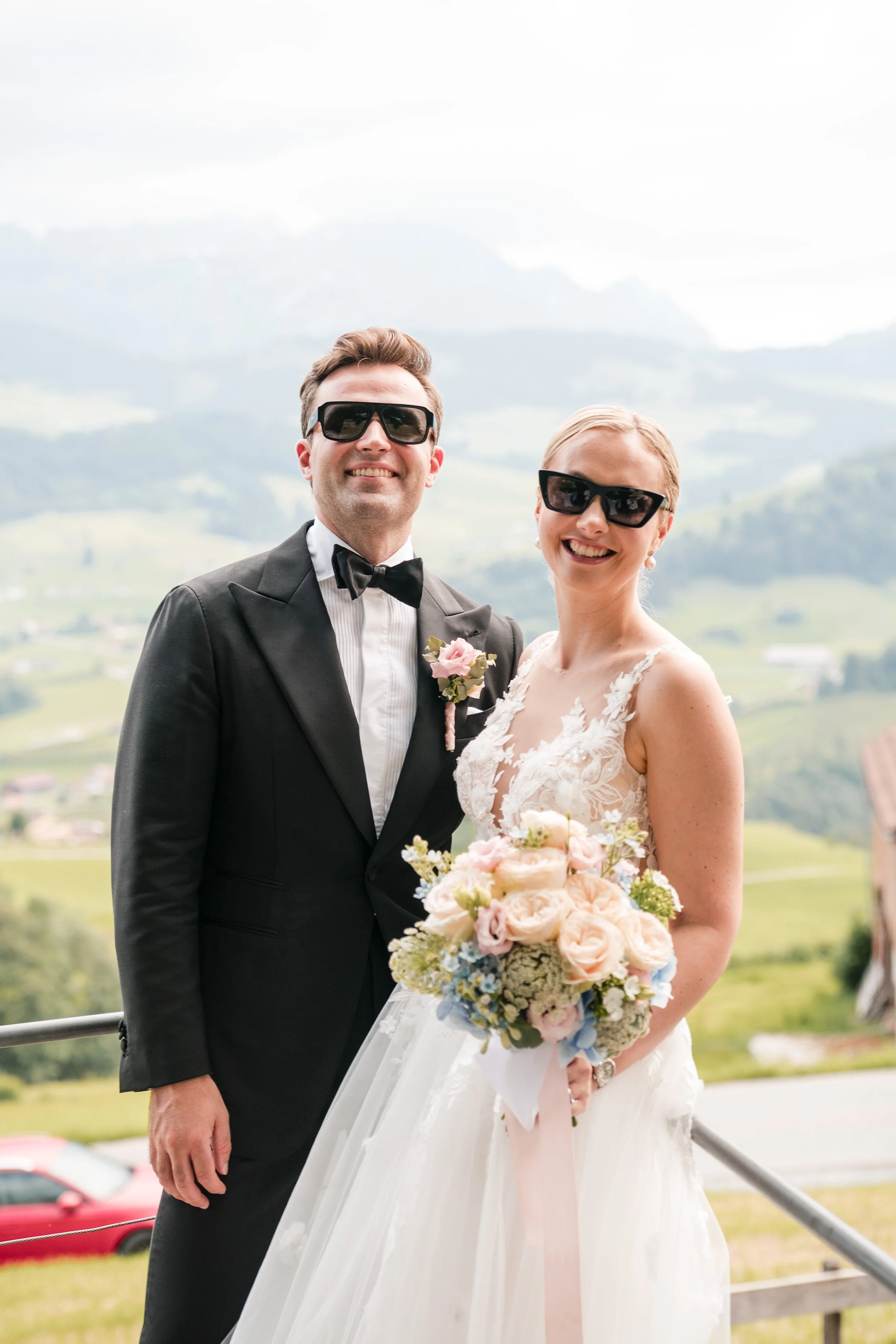 Ein lachendes Brautpaar in Hochzeitskleidung, beide tragen Sonnenbrillen, im Hintergrund eine bergige Landschaft.