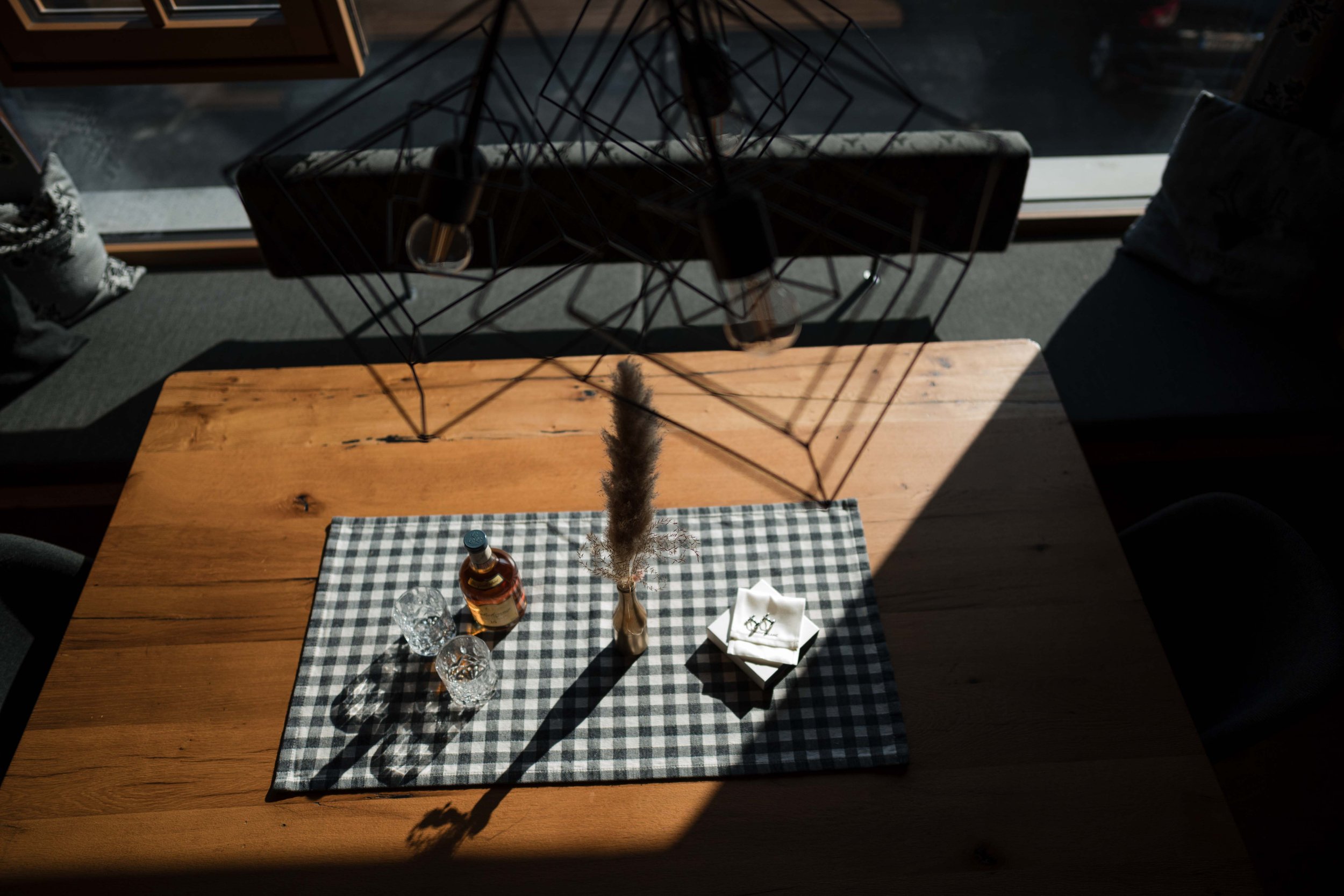 Ein Holztisch mit einer schwarzen und weißen Karo-Tischdecke, auf der eine Vase mit einer Tapfendeko, zwei Gläser, eine Flasche, und eine kleine weiße Schale mit Besteck stehen. Über dem Tisch hängt eine moderne schwarze Metallleuchte, und im Hinterg