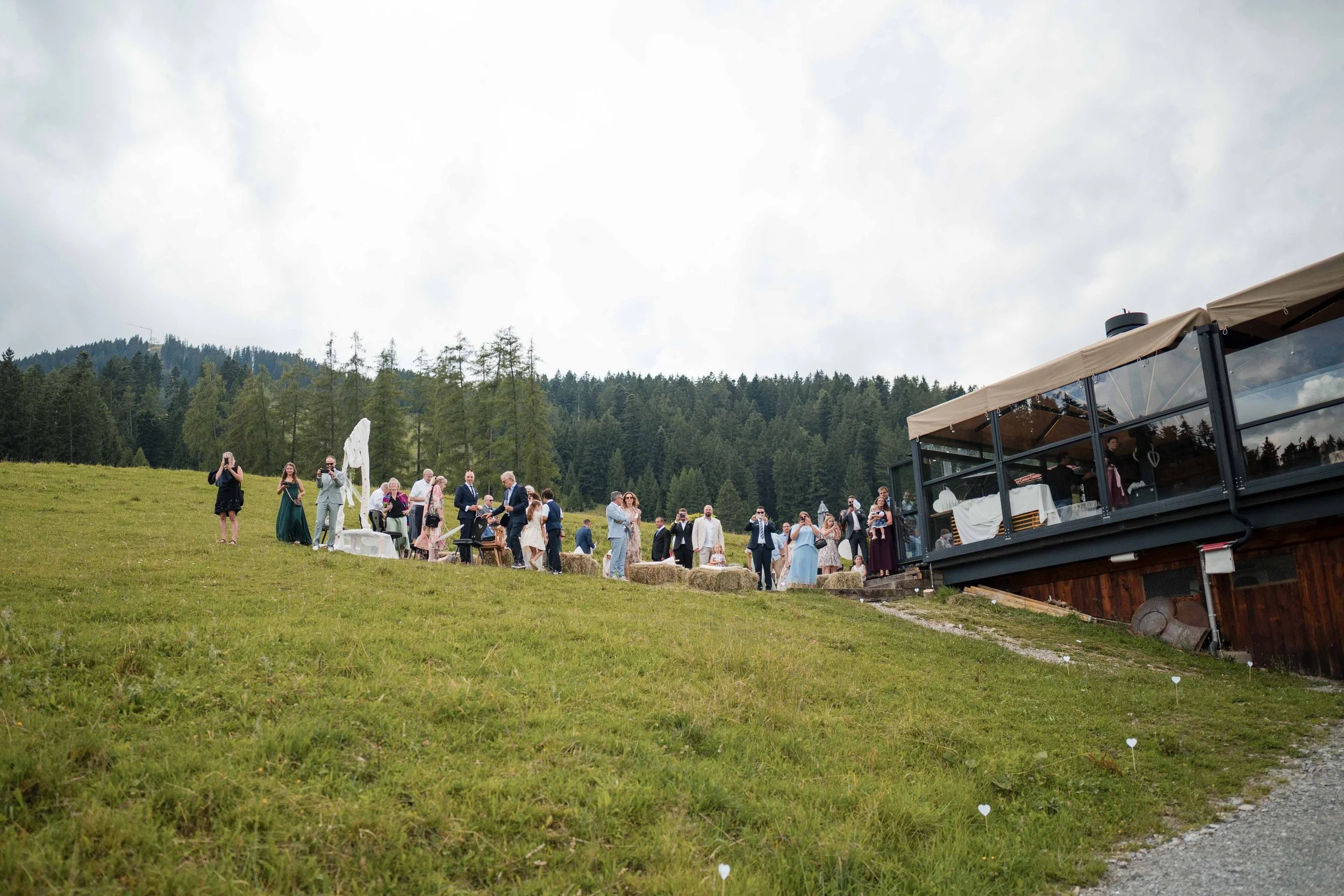 Menschen feiern auf einer Wiese im Freien, umgeben von Bäumen, bei einem eventuellen Hochzeitsfest, mit einem weißen Standbild und Strohballen, im Hintergrund ein Gebäude mit Terrasse.