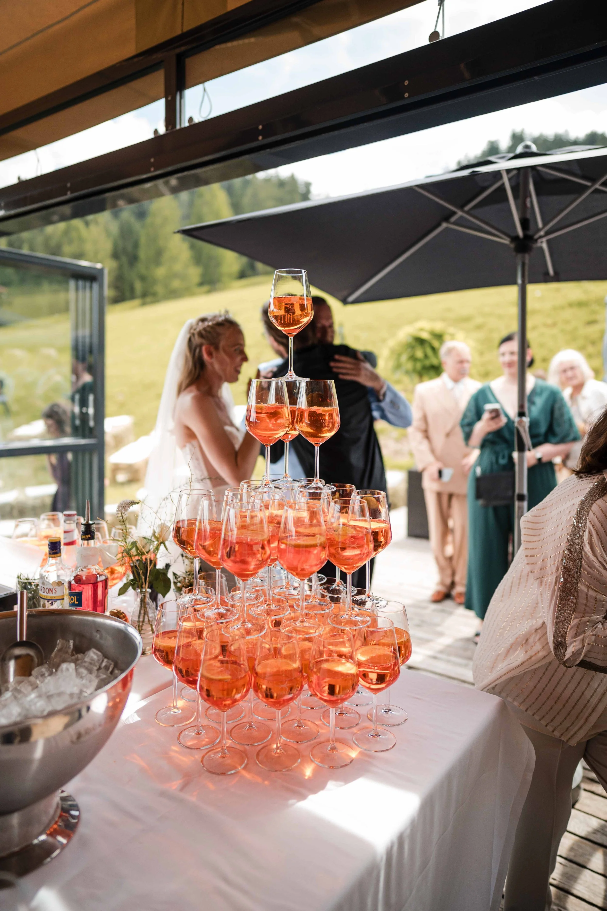 Hochzeitsparty im Freien mit Sektgläsern, die auf einem Tisch gestapelt sind, im Hintergrund embrace und Gäste bei einem Hochzeitsempfang bei sonnigem Wetter.