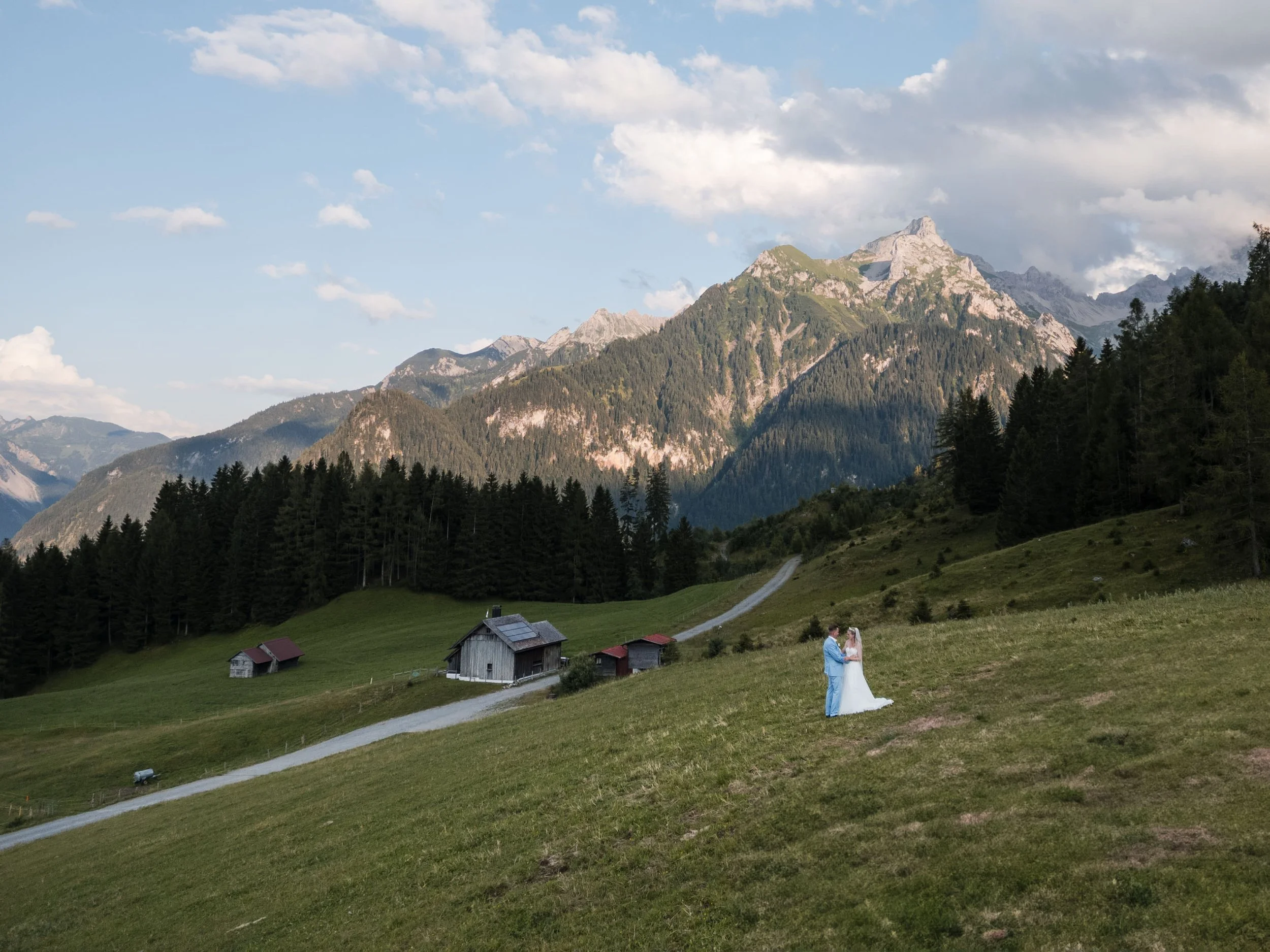 Ein Paar in Hochzeitskleidung steht auf einer grünen Wiese mit Bergkulisse. Es gibt einige kleine Häuser und einen Weg im Hintergrund.