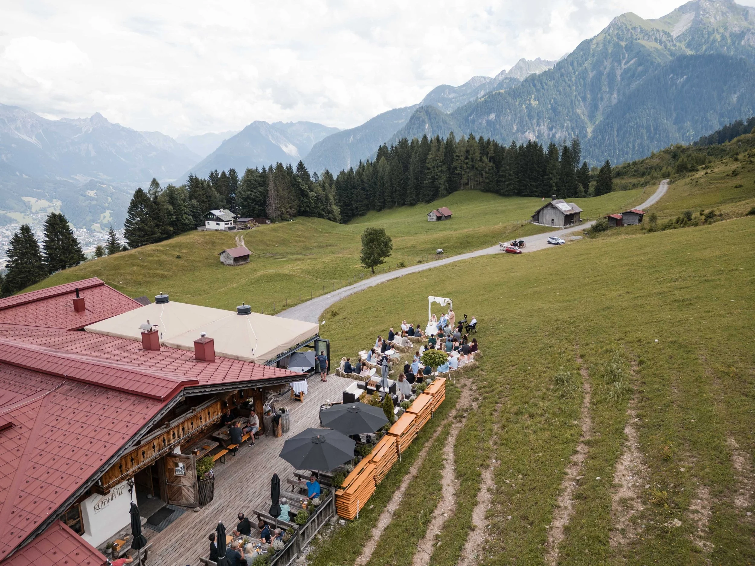 Eine Outdoor-Hochzeit im Grünen mit vielen Gästen auf einer Terrasse vor einem Bauernhaus, inmitten einer bergigen Landschaft.