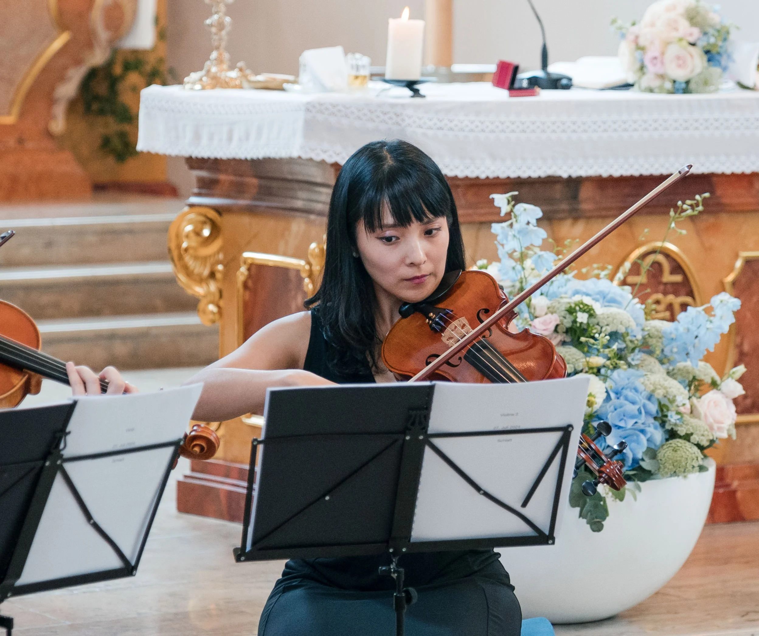 Eine junge Frau spielt Geige bei einem Konzert, umgeben von Musiknotenständern und Blumendekorationen im Hintergrund.