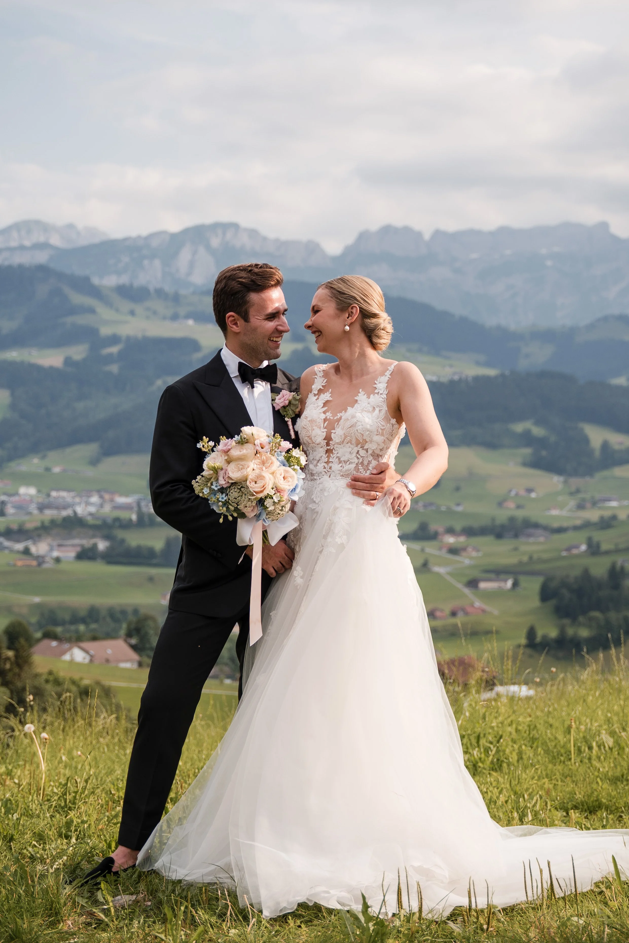 Ein Brautpaar steht auf einer Wiese vor den Bergen, das Brautpaar lächelt sich an, die Braut trägt ein weißes Hochzeitskleid mit Spitzen-Details, der Bräutigam einen schwarzen Anzug mit Fliege, beide sehen glücklich aus, die Braut hält einen Blumenst