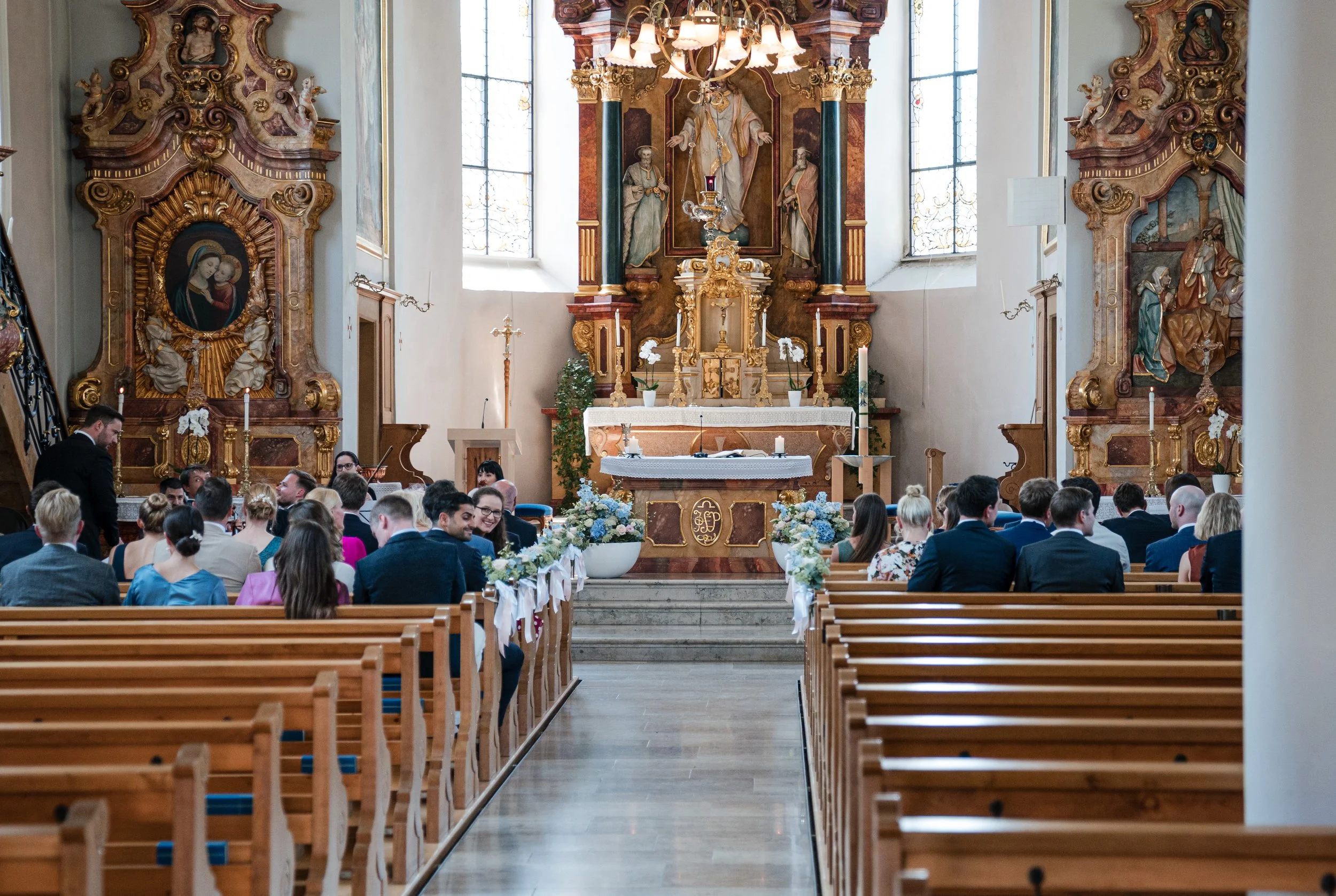 Innenansicht einer Kirche während einer Hochzeitszeremonie, mit anwesenden Gästen, einem Altar, religiösen Kunstwerken und Kerzen.