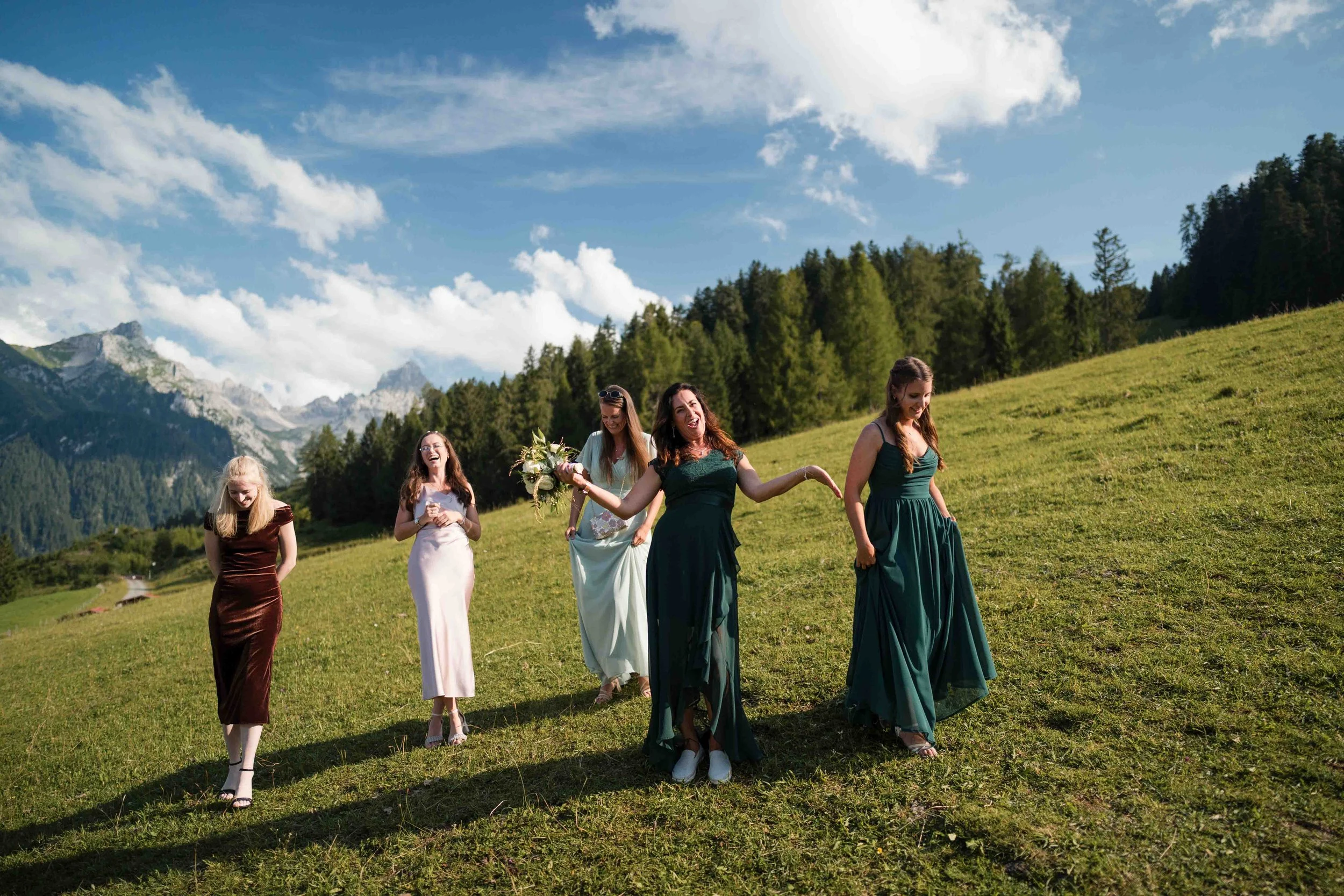 Fünf Frauen in eleganten Kleidern laufen auf einer grünen Wiese mit Bergen im Hintergrund, sonniger Himmel mit Wolken, Wohlfühlatmosphäre.