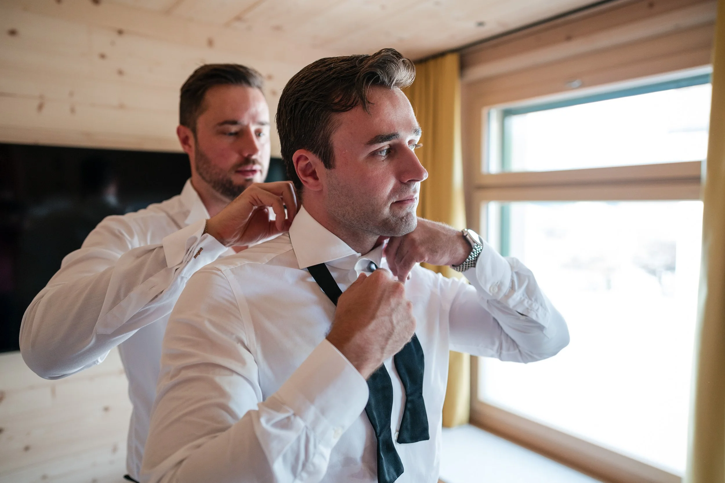 Zwei Männer in weißen Hemden, einer hilft dem anderen, seine Krawatte zu binden, in einem Raum mit Holzdecke und großen Fenstern, gelben Vorhängen.