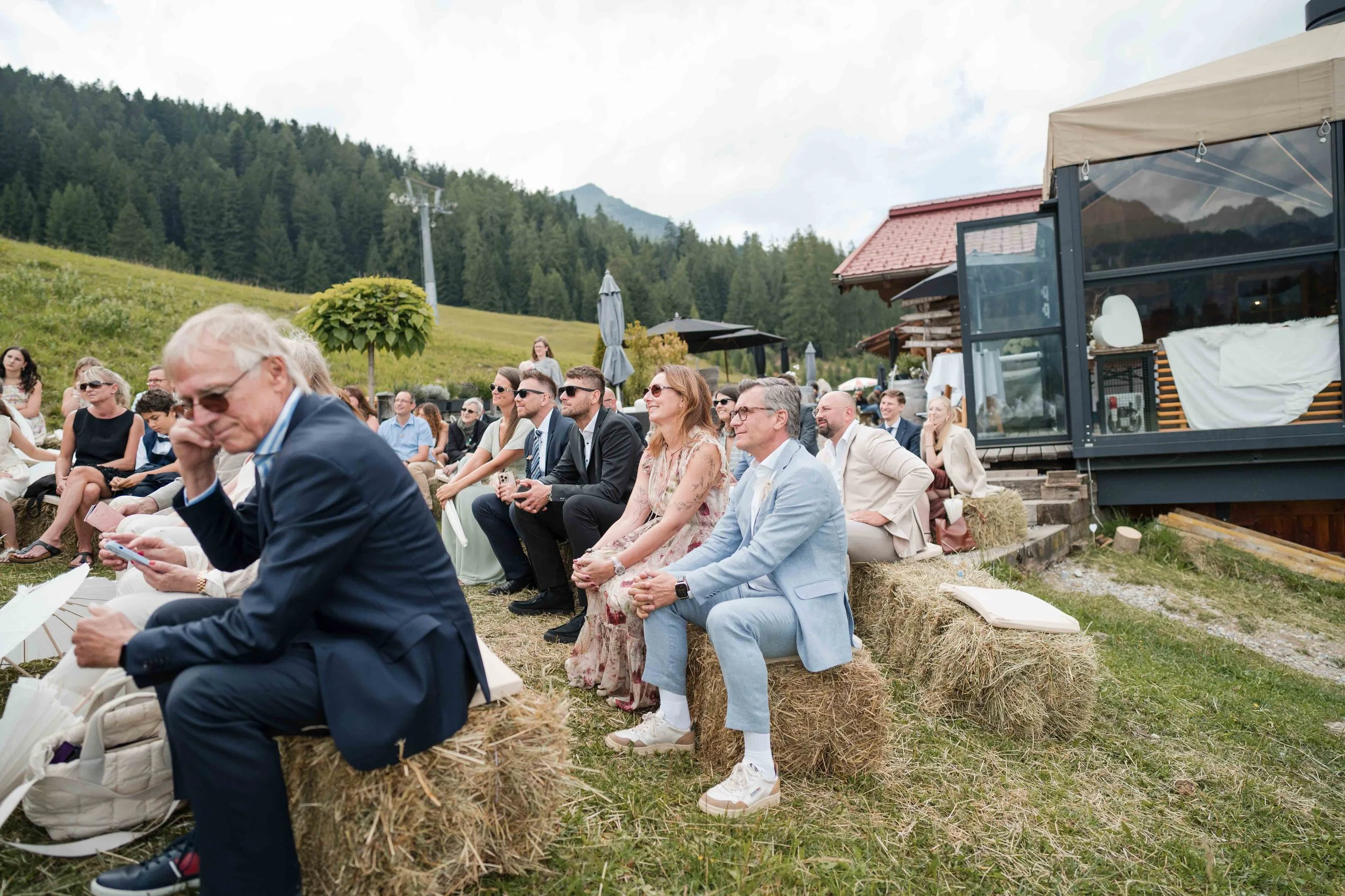 Menschen sitzen im Freien auf Heuballen bei einer Veranstaltung, umgeben von grünen Hügeln und Bergen, mit einem Zelt im Hintergrund.