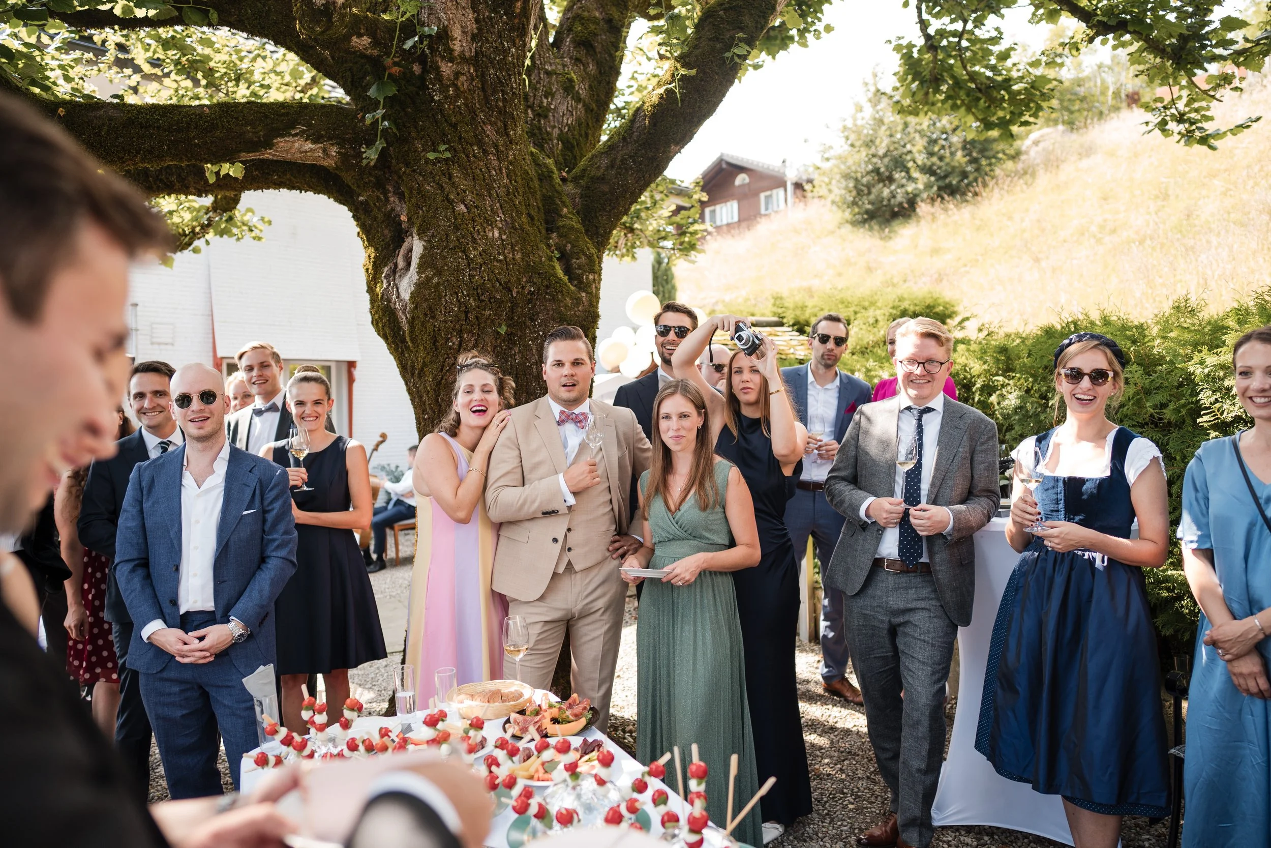 Eine Gruppe von Menschen bei einer outdoor Feier, vermutlich Hochzeit, unter einem Baum, die sich um einen Tisch mit Kuchen und Snacks versammelt, lachend und fröhlich.