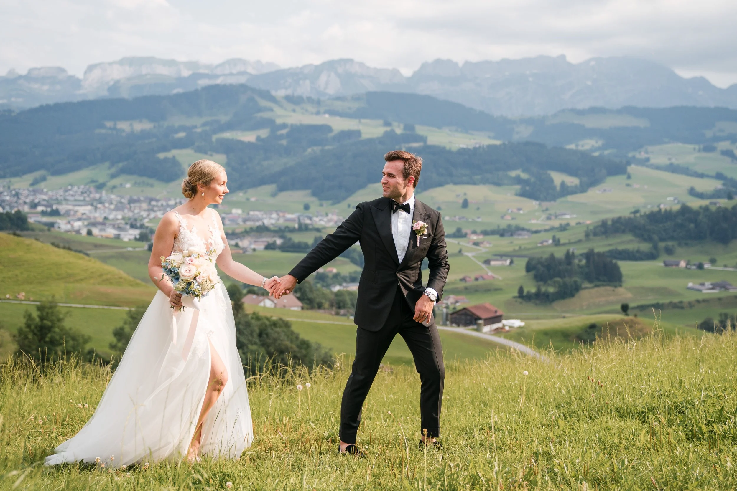 Ein Brautpaar hält sich an einem Hügel in einer grünen Landschaft mit Bergen im Hintergrund. Die Braut trägt ein weißes Hochzeitskleid und hält einen Blumenstrauß, der Bräutigam trägt einen schwarzen Anzug und eine Fliege.