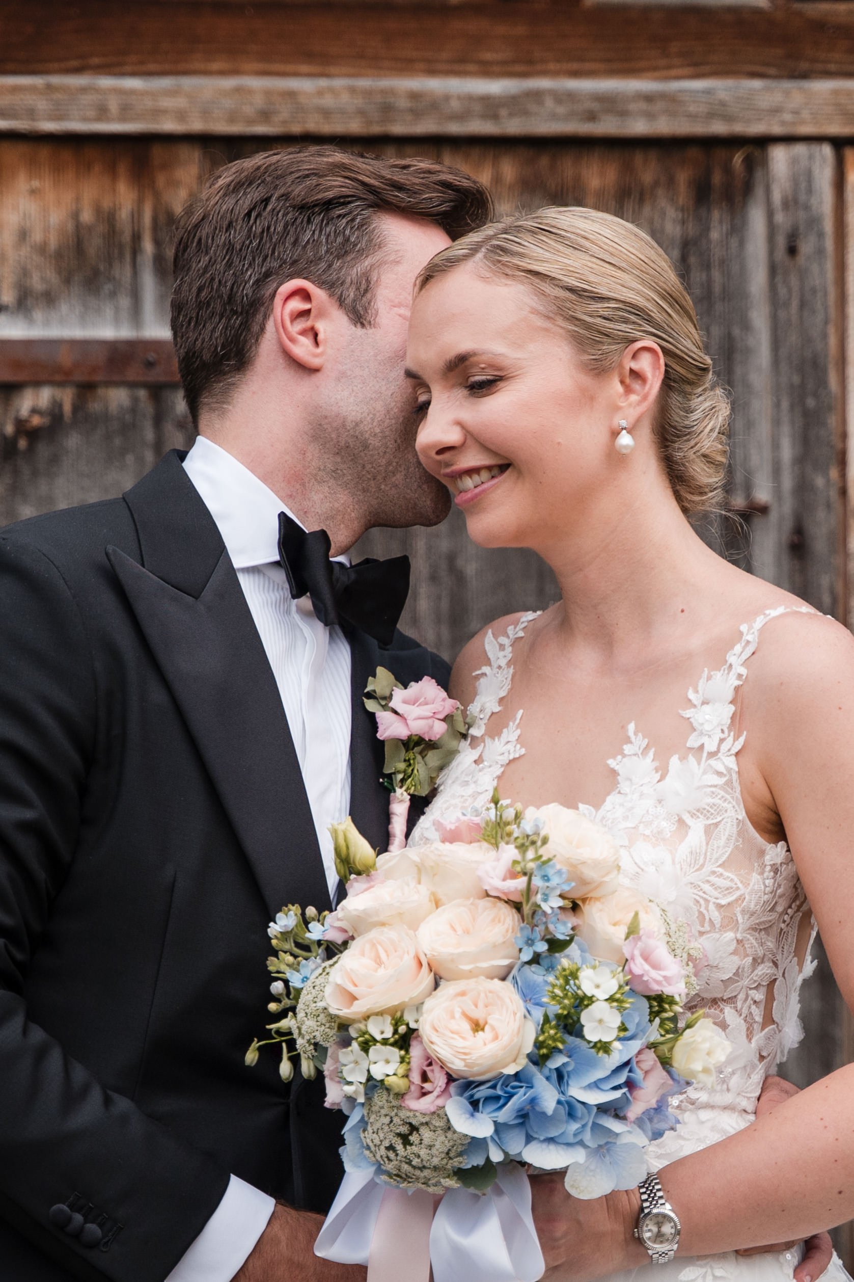 Ein Brautpaar bei ihrer Hochzeit. Sie lächeln, der Bräutigam trägt einen schwarzen Anzug und Fliege, die Braut trägt ein weißes Hochzeitskleid und hält einen Blumenstrauß.