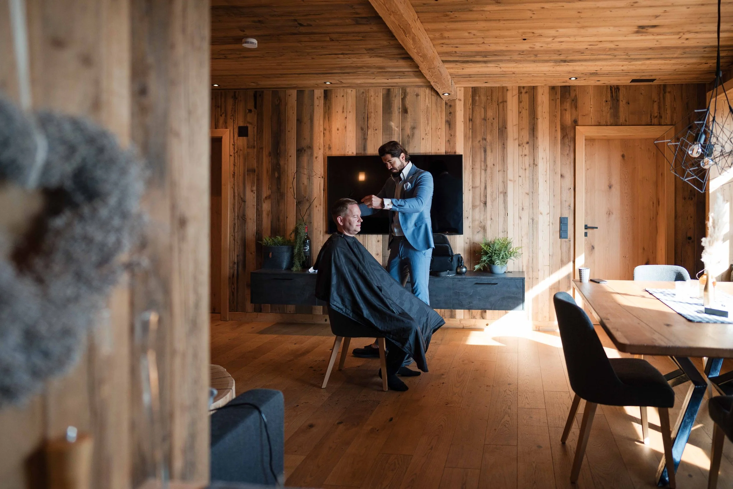 Ein Mann in einem blauen Anzug bekommt einen Haarschnitt in einem Holzinterieur. Ein Friseur schneidet ihm die Haare, während der Mann auf einem Stuhl sitzt.