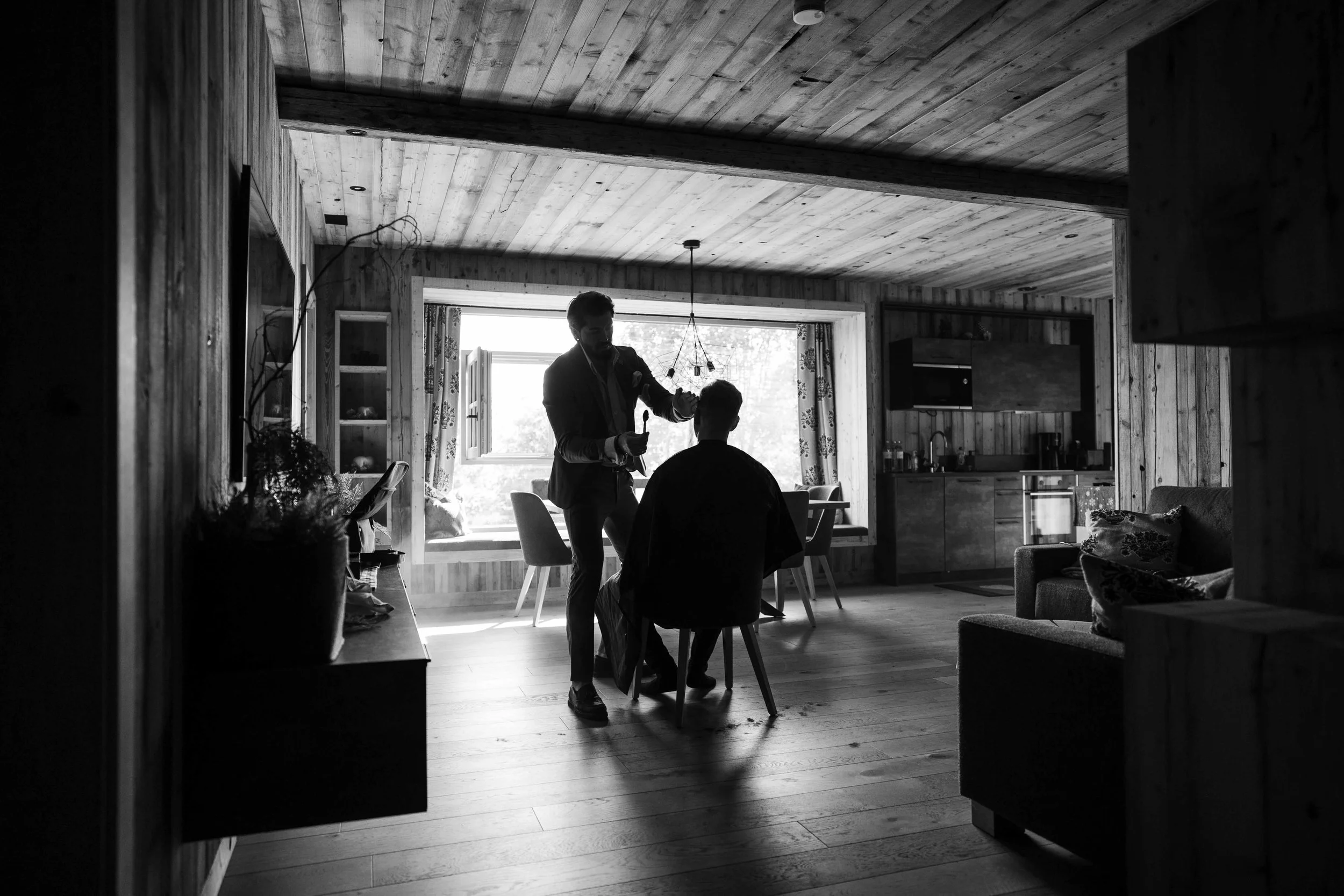 Zwei Männer in einem rustikalen, holzverkleideten Raum, einer steht und stylt den anderen, der auf einem Stuhl sitzt, vor einem großen Fenster.