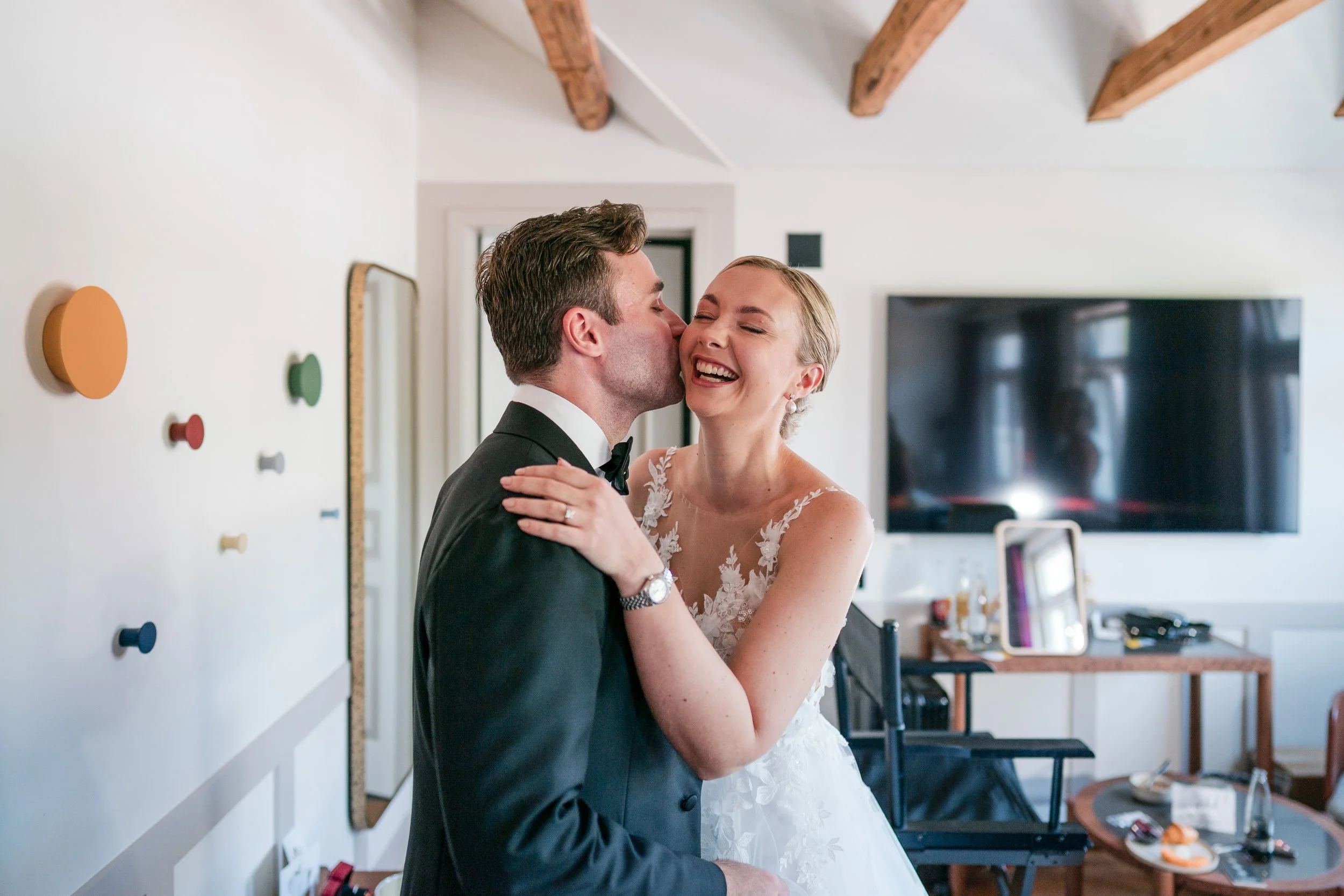 Ein junges Brautpaar, das sich bei ihrer Hochzeit küsst, in einem modernen Raum mit bunten Wanddekorationen und einem Fernseher im Hintergrund.