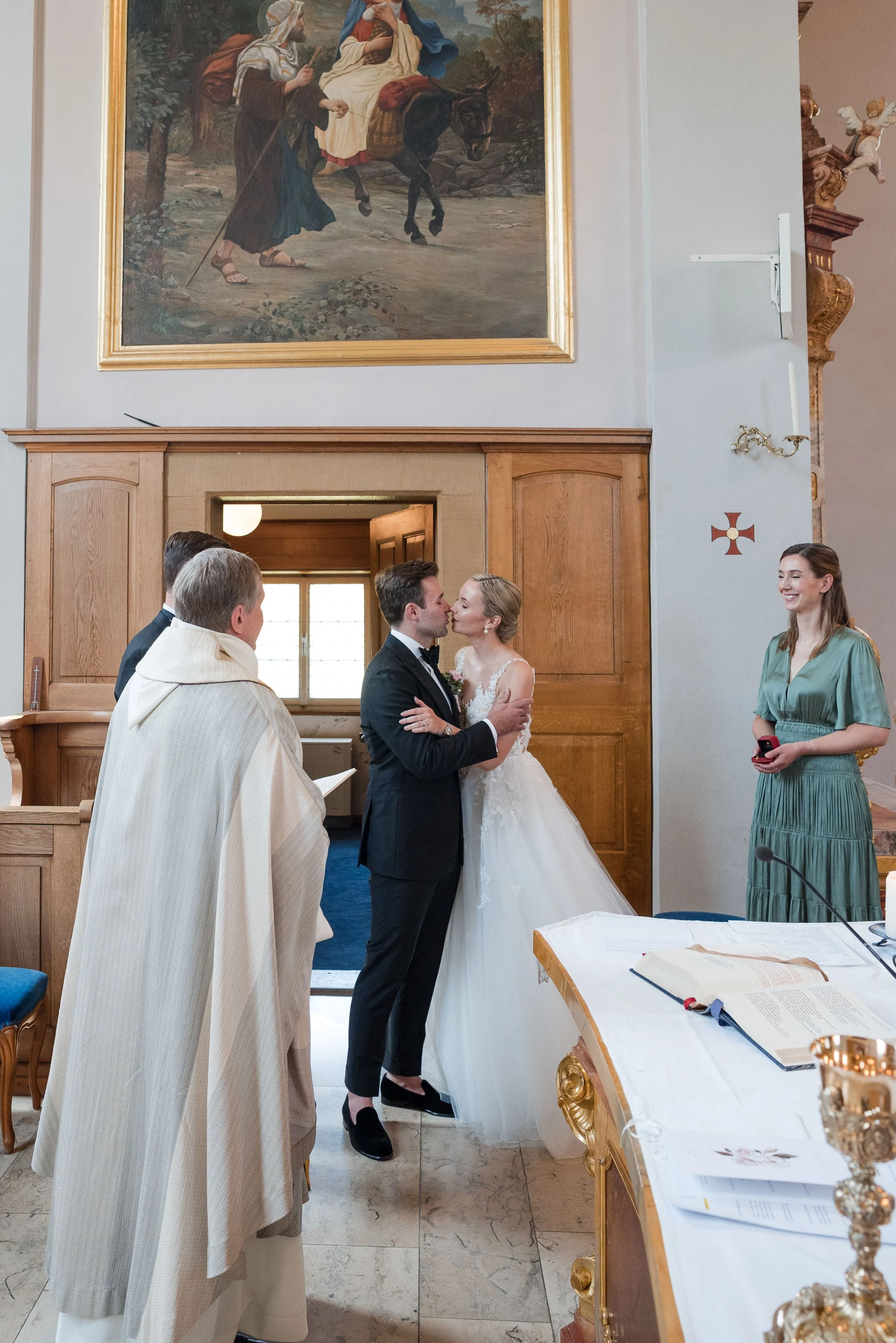 Ein Hochzeitspaar tauscht einen Kuss während der Trauung in einer Kirche, umgeben von Trauzeugen und einem Priester, mit einem großen Gemälde an der Wand im Hintergrund.