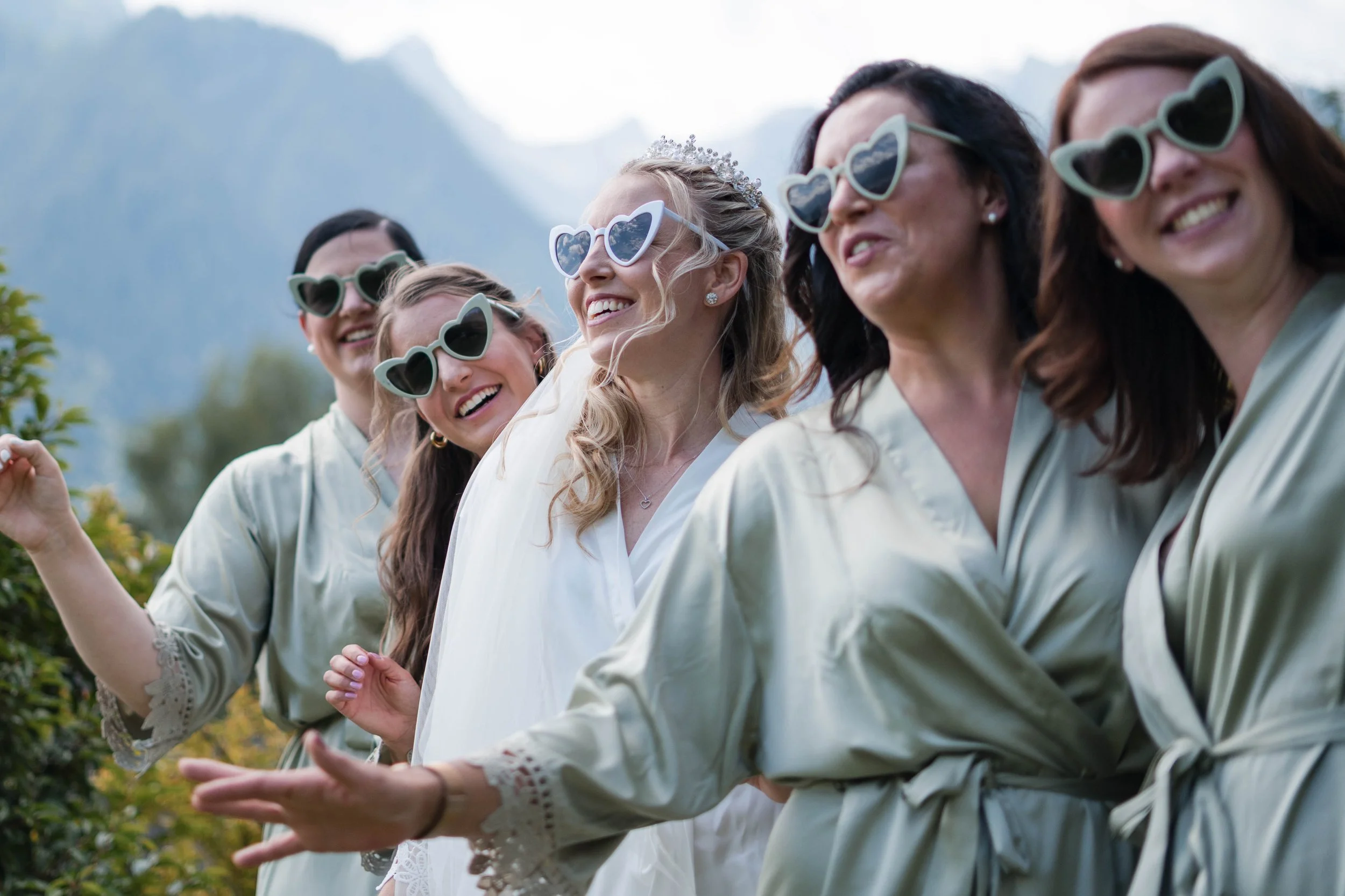 Fünf Frauen in Braut- und Brautjungfernkleidern, lachen und posieren mit Herz-Sonnenbrillen im Freien vor Bergen.