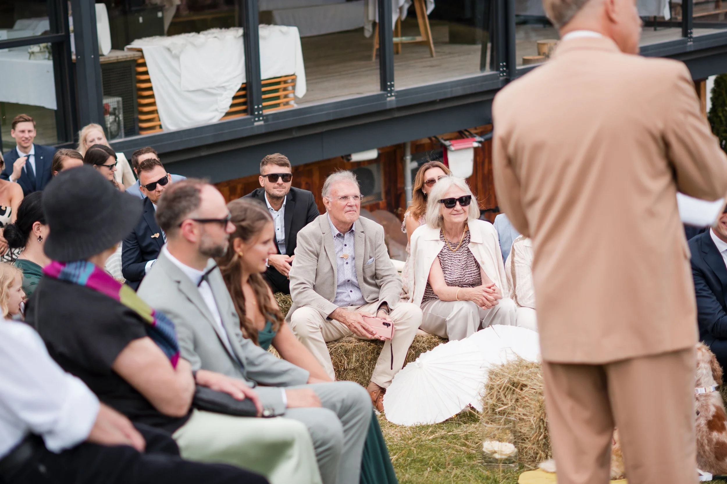 Menschen sitzen bei einer Outdoor-Veranstaltung in einer Reihe. Einige tragen Sonnenbrillen und formelle Kleidung, während ein paar ältere Damen mit weißem Haar und Sonnenbrillen im Vordergrund sitzen. Im Vordergrund ist eine Person in einem hellbrau