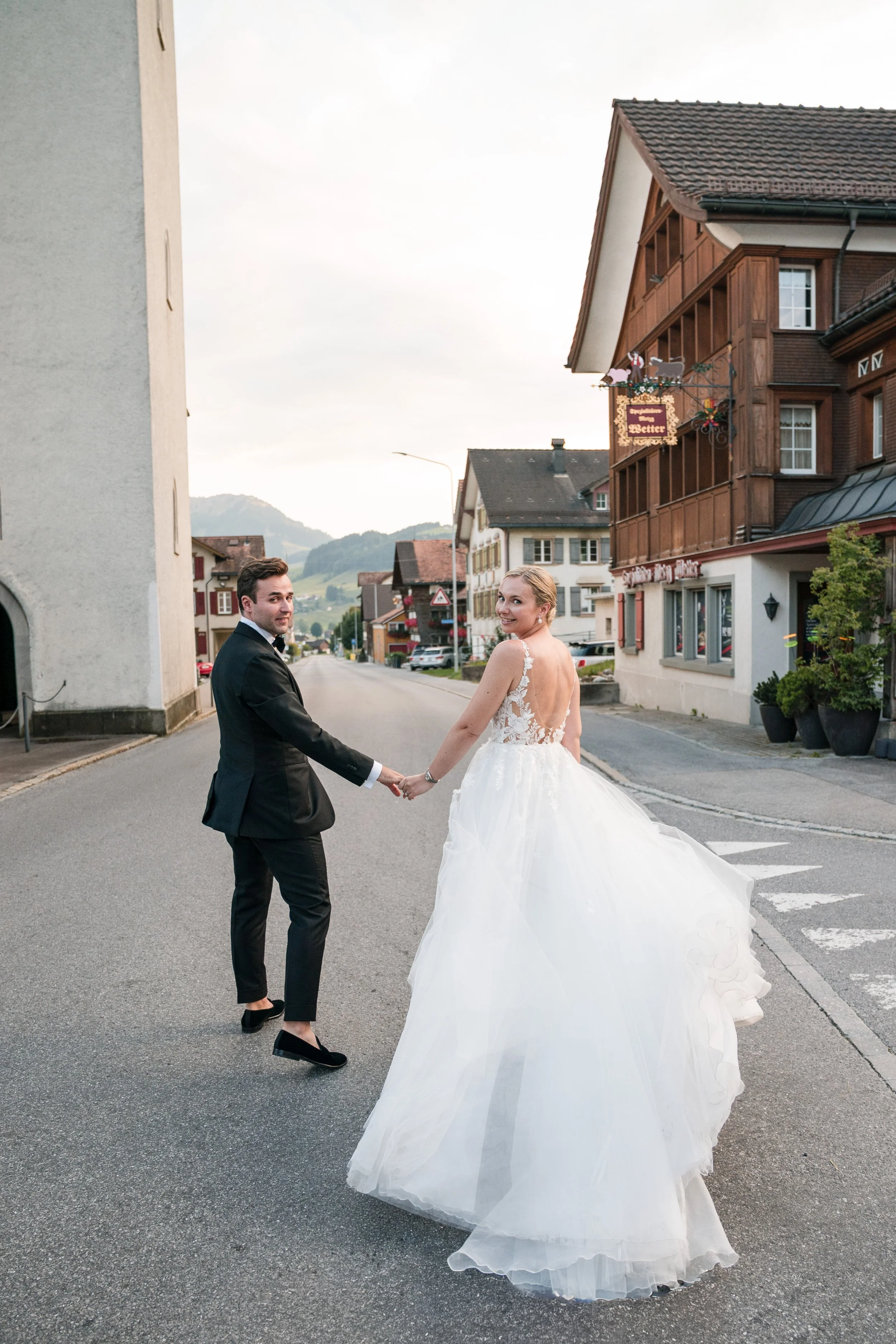 Ein Brautpaar hält sich an einer ruhigen Straße in einem kleinen Dorf in der Schweiz bei Sonnenuntergang, im Hintergrund Berge, die Frau trägt ein weißes Brautkleid, der Mann einen schwarzen Anzug.