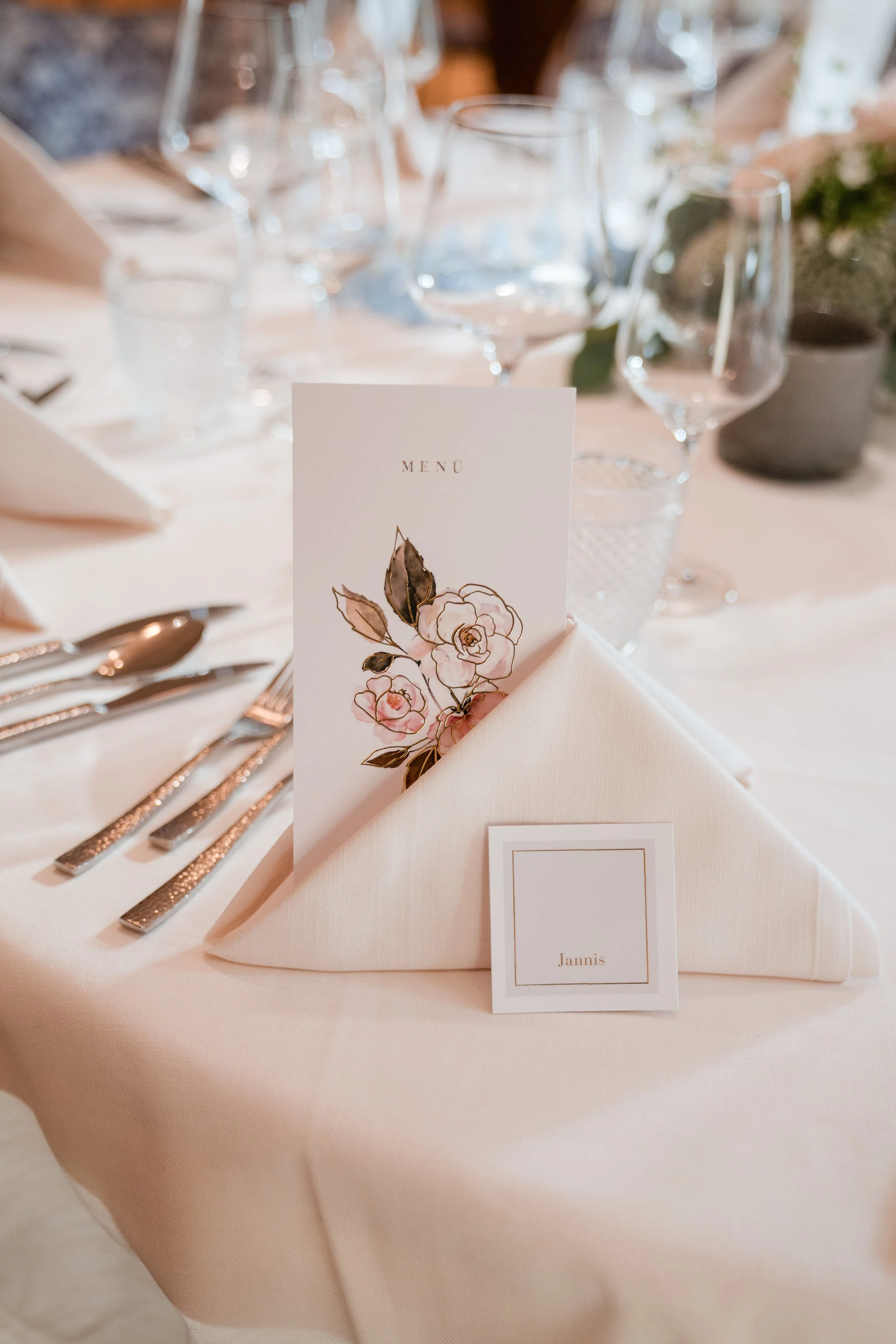 Eleganter Tisch bei einer Veranstaltung, mit weißen Tafel, Weingläsern, Besteck, einem Menükarte mit Blumenmotiv, und einem Namensschild mit der Aufschrift 'Jannis'.