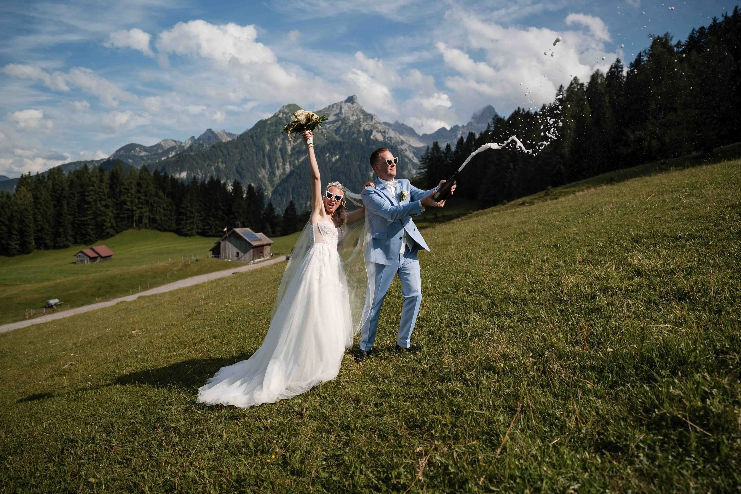 Ein Brautpaar feiert Hochzeit im Grünen, sie trägt ein weißes Brautkleid und er einen hellblauen Anzug, sie lachen und halten eine Flasche Champagner, während sie auf einer Wiese vor einer Bergkulisse stehen.