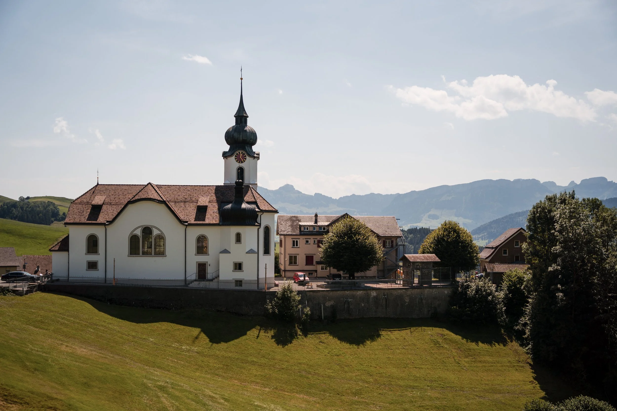 Eine weiße Kirche mit dunklem Turm vor einer ländlichen Landschaft mit Bergen im Hintergrund, Bäumen und einigen Häusern.