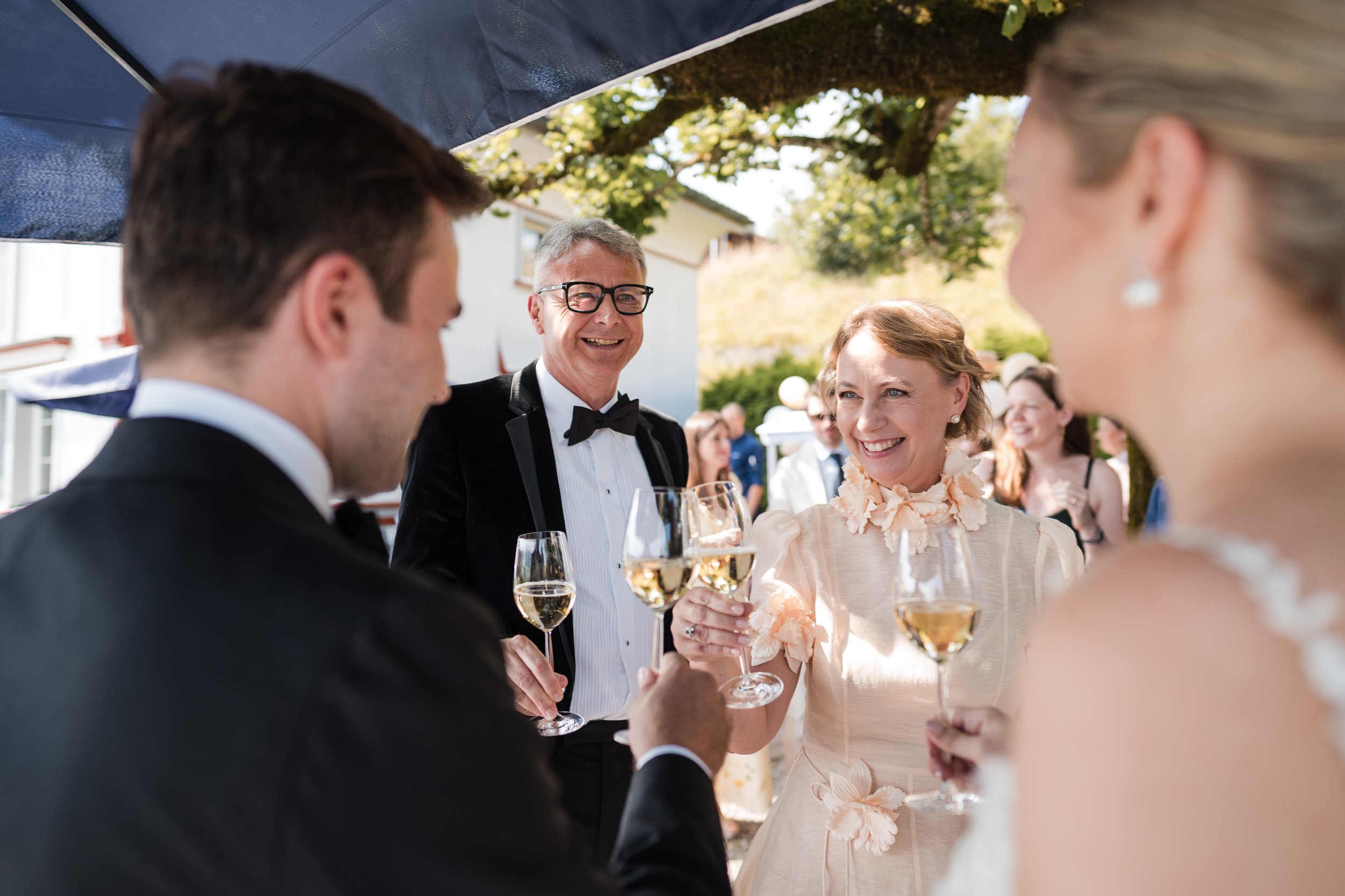 Menschen bei einer Hochzeitsfeier, die Gläser mit Sekt trinken, im Freien unter Sonnenschirmen.