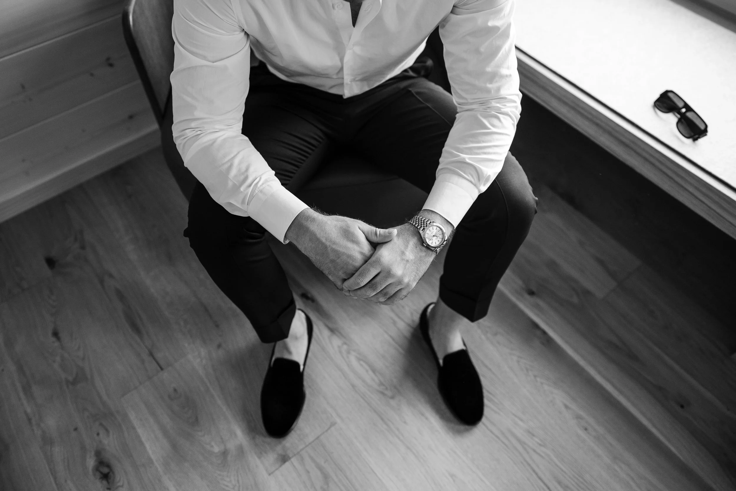 Ein Mann sitzt auf einem Stuhl, trägt ein weißes Hemd, schwarze Hosen und Loafers, mit einer Uhr am Handgelenk, auf einer Holzfußboden. Er hat die Hände vor dem Körper verschränkt.