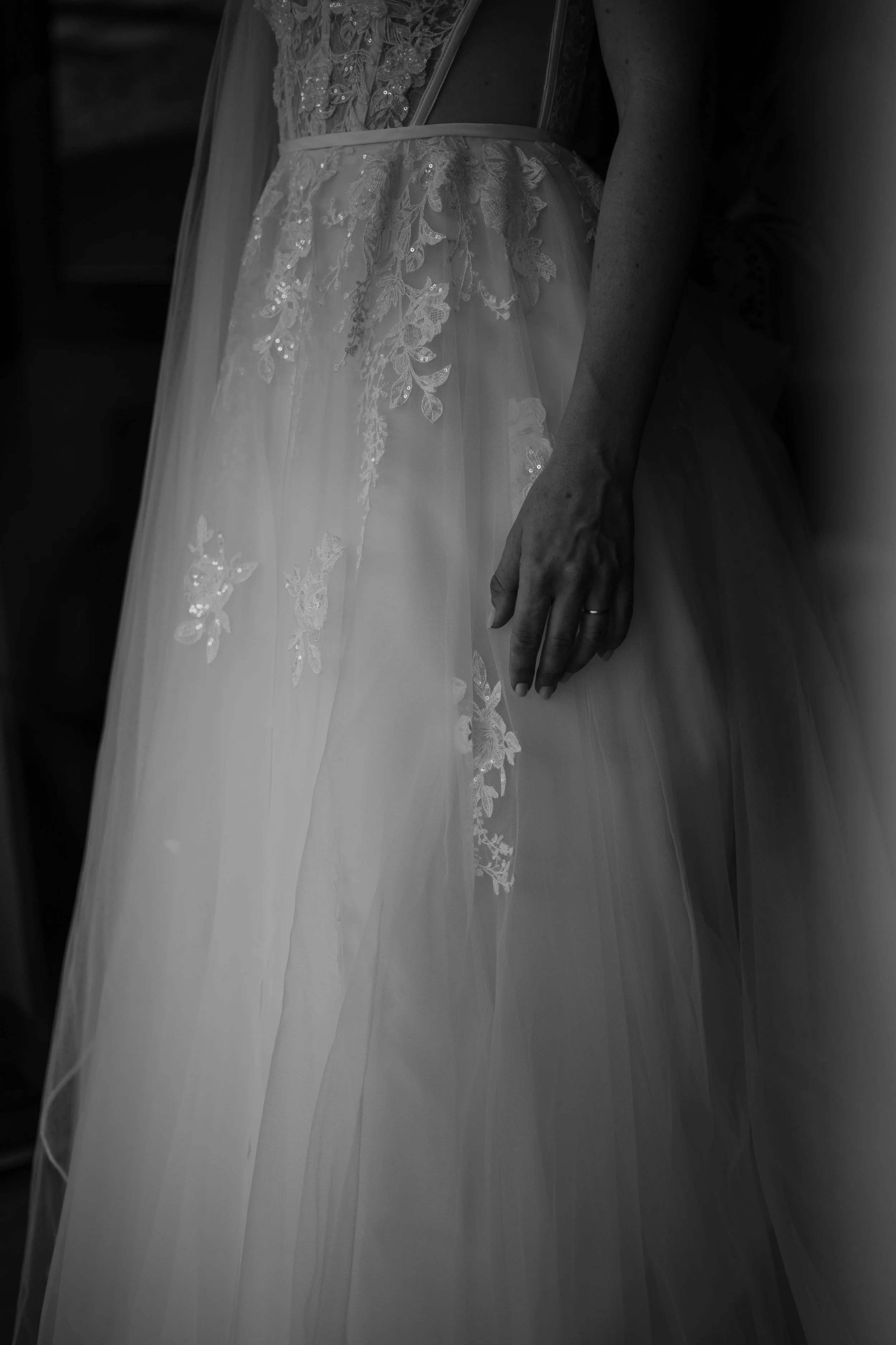 Schwarz-weiß-Fotografie einer Braut in Hochzeitskleid mit floral besticktem Tüll, deren Hand am Rock hängt.