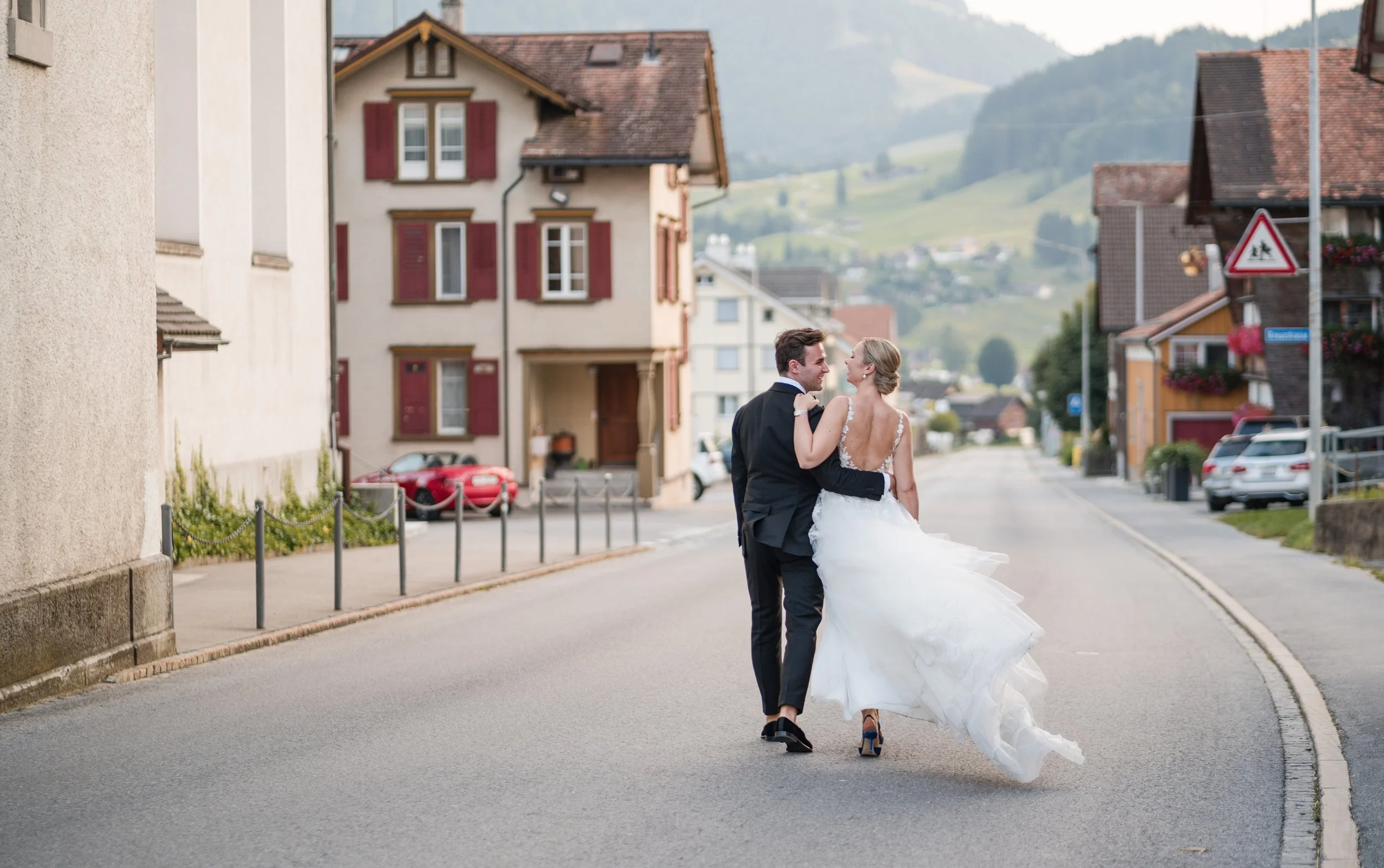 Ein Brautpaar läuft Hand in Hand durch eine ruhige Straße in einem Dorf, umgeben von traditionellen Häusern und grünen Hügeln im Hintergrund.
