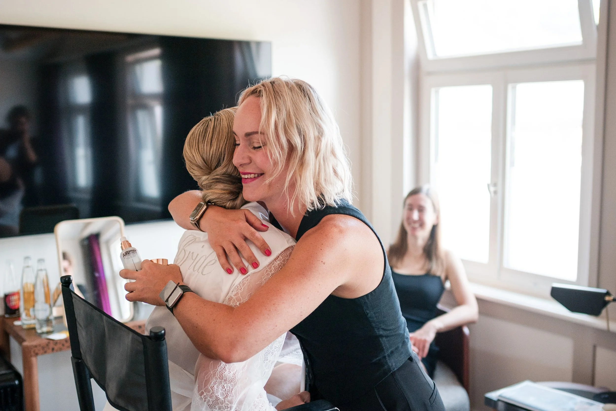Zwei Frauen umarmen sich herzlich im Zimmer, eine trägt ein Hochzeitskleid und die andere ein schwarzes Kleid, während eine dritte Person im Hintergrund lächelt.