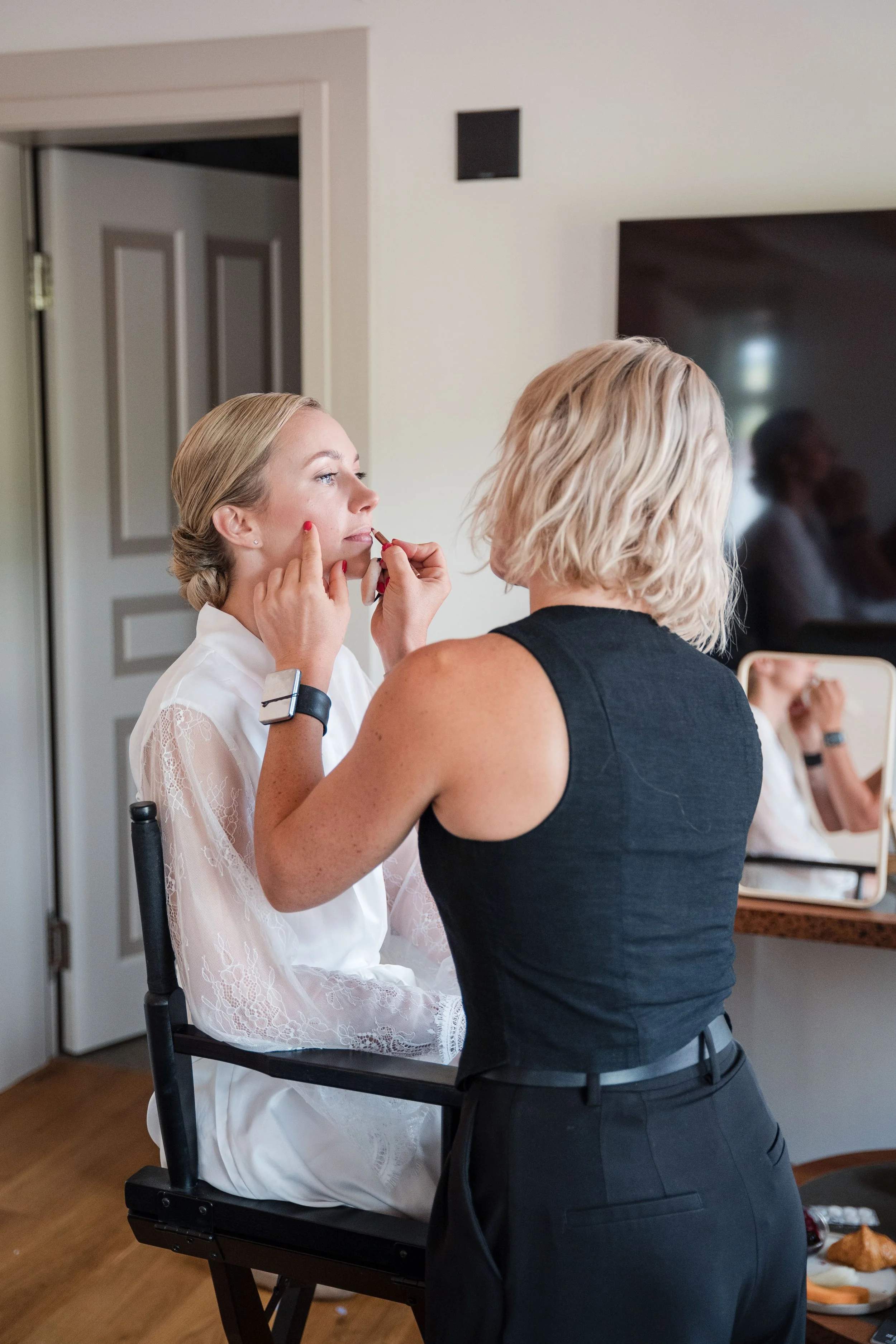 Eine Frau wird für eine Hochzeit geschminkt, während eine makeup-artist sie dabei berät, im Hintergrund ein Spiegel mit einem reflektierten Selbstporträt und eine weitere Person sind sichtbar.