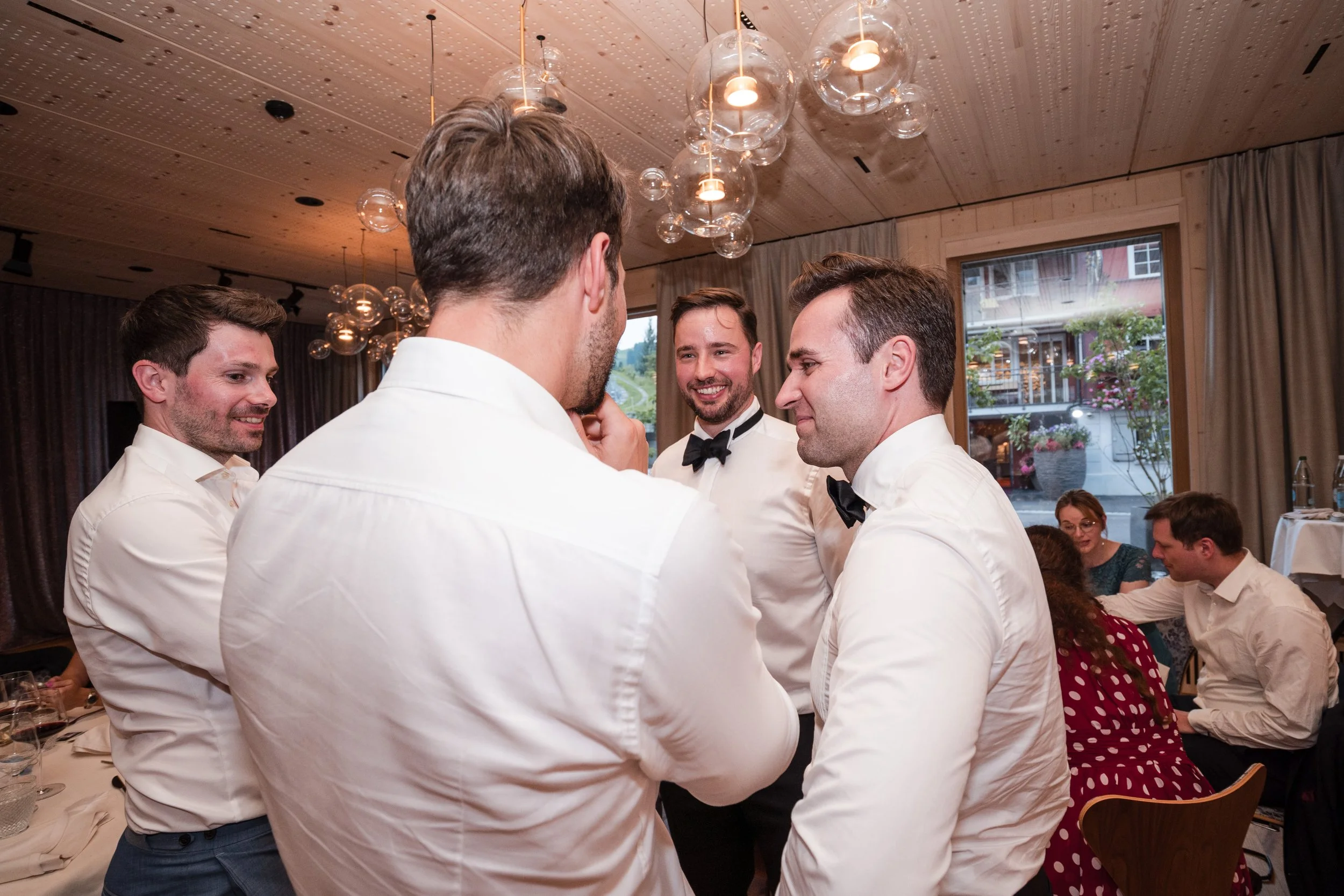 Fünf Männer in weißem Hemd, einige mit Fliegen, haben eine Unterhaltung bei einem festlichen Anlass in einem großzügigen Raum mit Holzwanddecke und großen Fenstern mit Blick auf eine Außenanlage