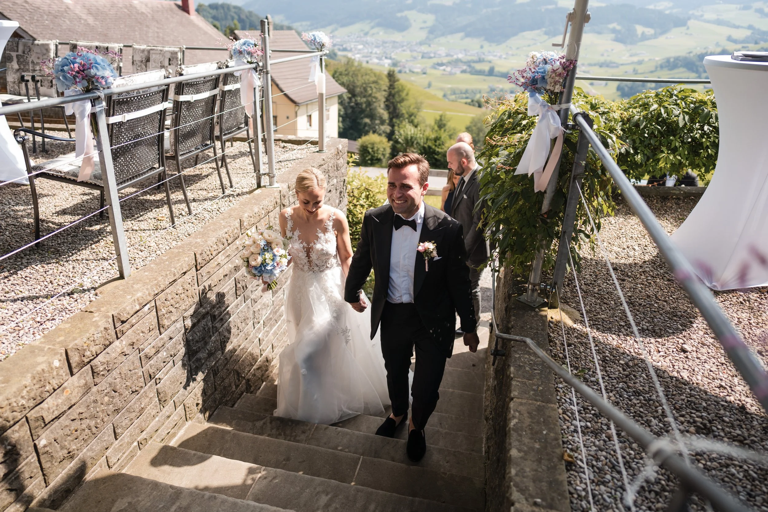 Ein Brautpaar steigt die Treppen eines Hauses hinauf, die Braut trägt ein weißes Hochzeitskleid und hält einen Blumenstrauß, der Bräutigam trägt einen schwarzen Anzug mit Fliege. Im Hintergrund sind weitere Gäste und eine ländliche Landschaft mit Hüg