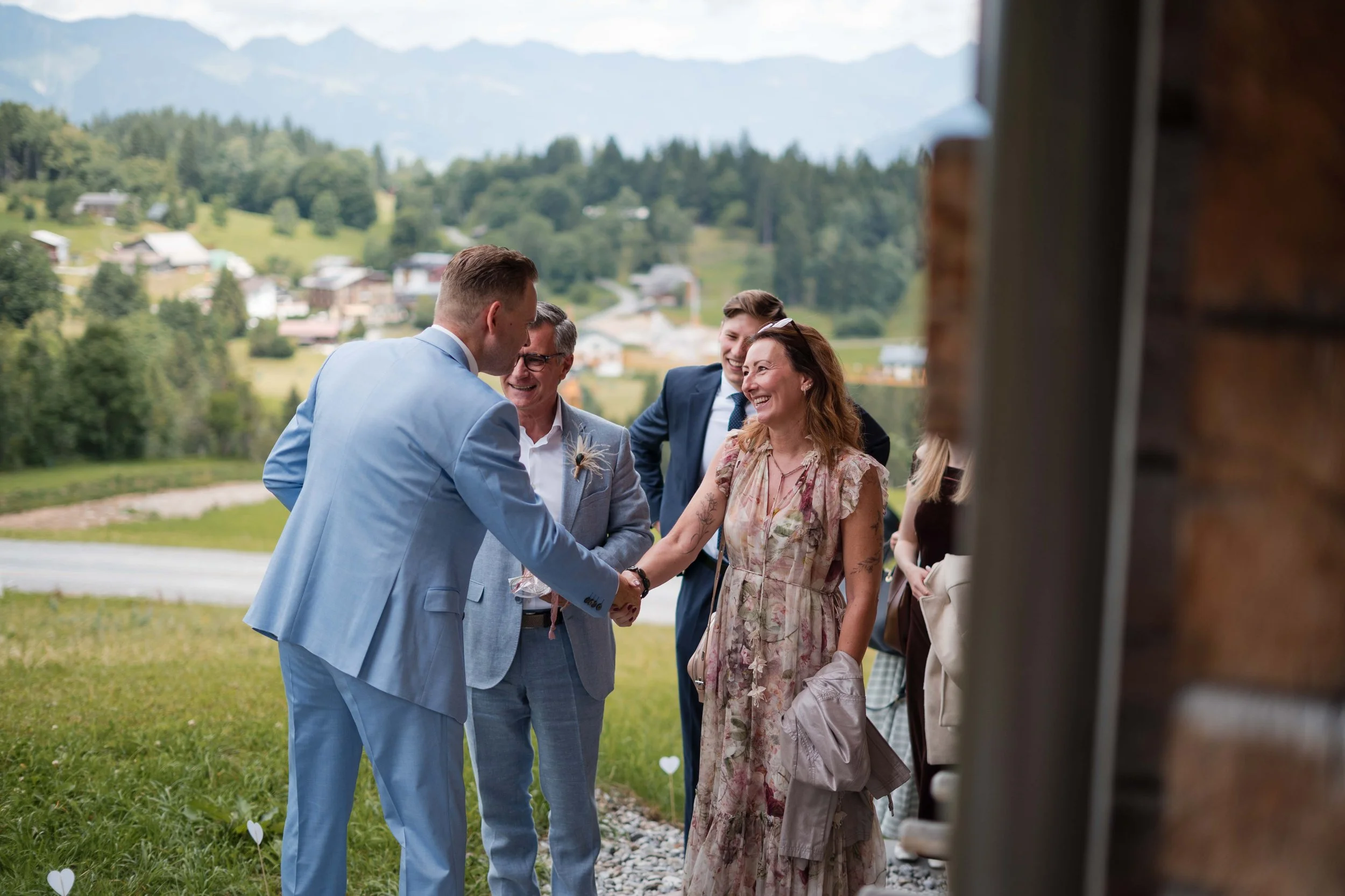 Hochzeit im Freien mit lachenden Gästen, die den Brautpaar begrüßen, in einer ländlichen Umgebung mit Hügeln und Häusern im Hintergrund.
