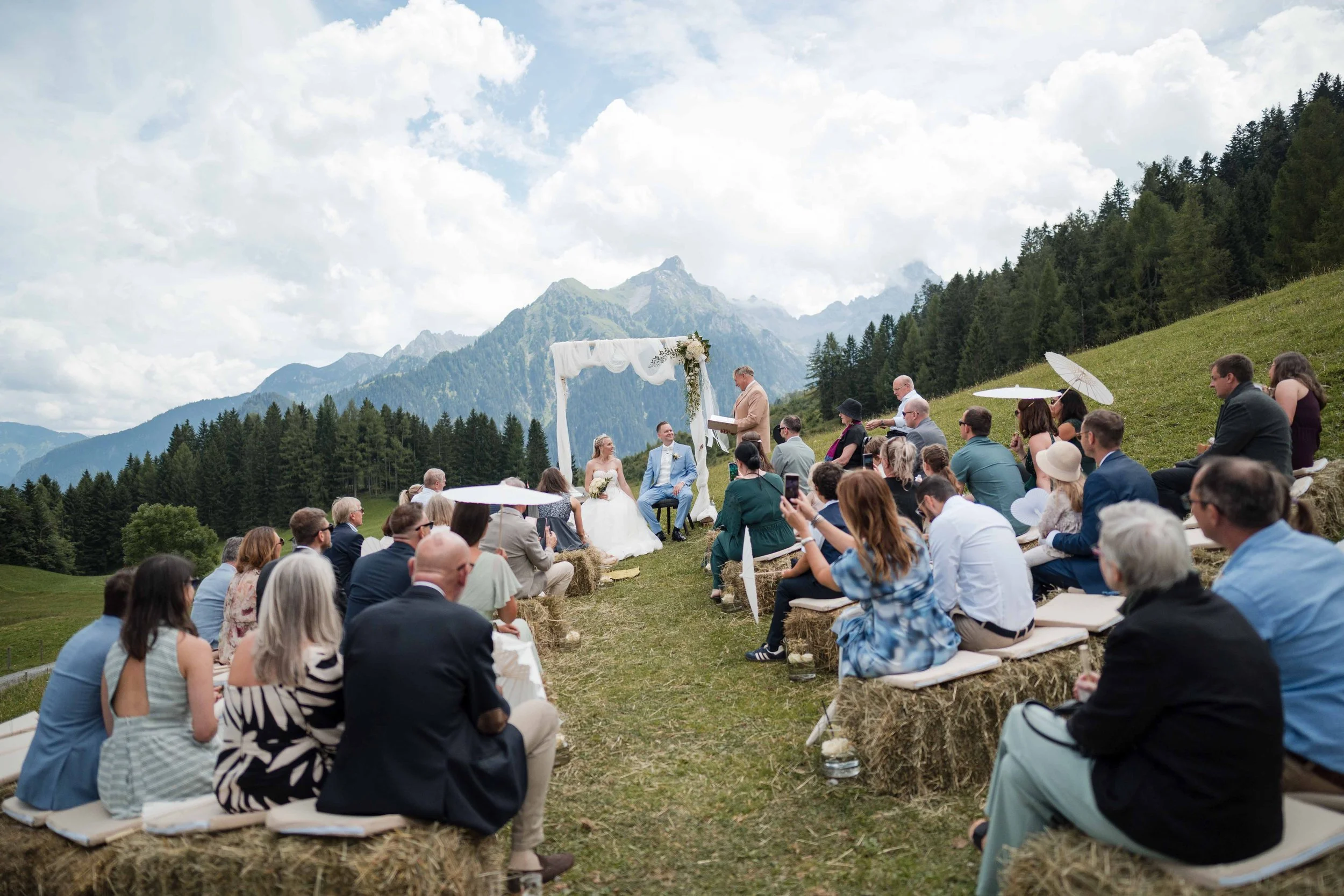 Eine Hochzeit im Freien in den Bergen mit Gästen, die auf Heuballen sitzen, ein Altar und eine zeremonielle Szene im Mittelpunkt，高 eine Landschaft mit hohen Bergen, Wäldern, einem blauen Himmel und Wolken im Hintergrund.
