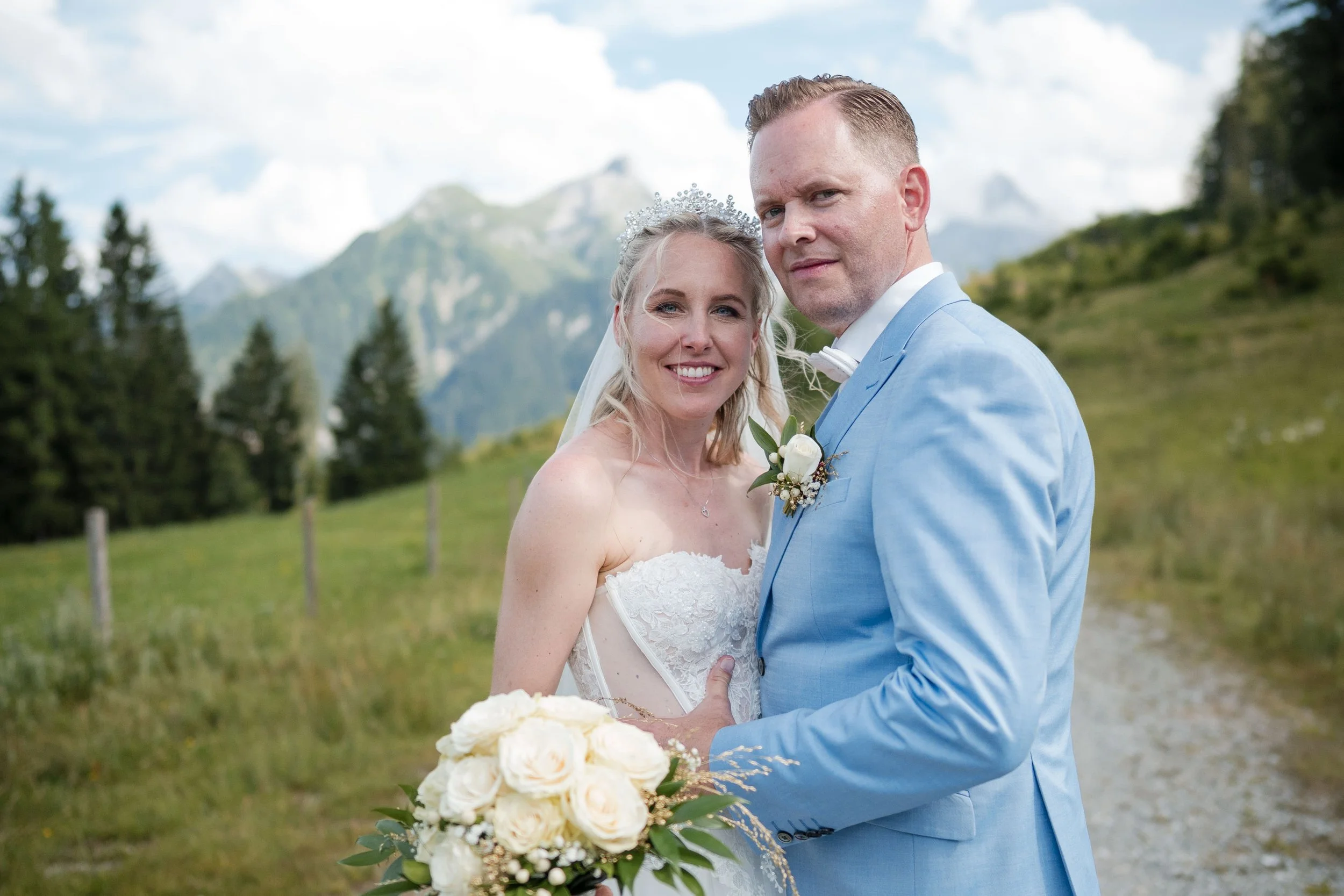 Ein Brautpaar steht im Freien in einer ländlichen Landschaft mit Bäumen und Bergen im Hintergrund, trägt Hochzeitskleidung und schaut in die Kamera, die Braut hält einen Blumenstrauß.