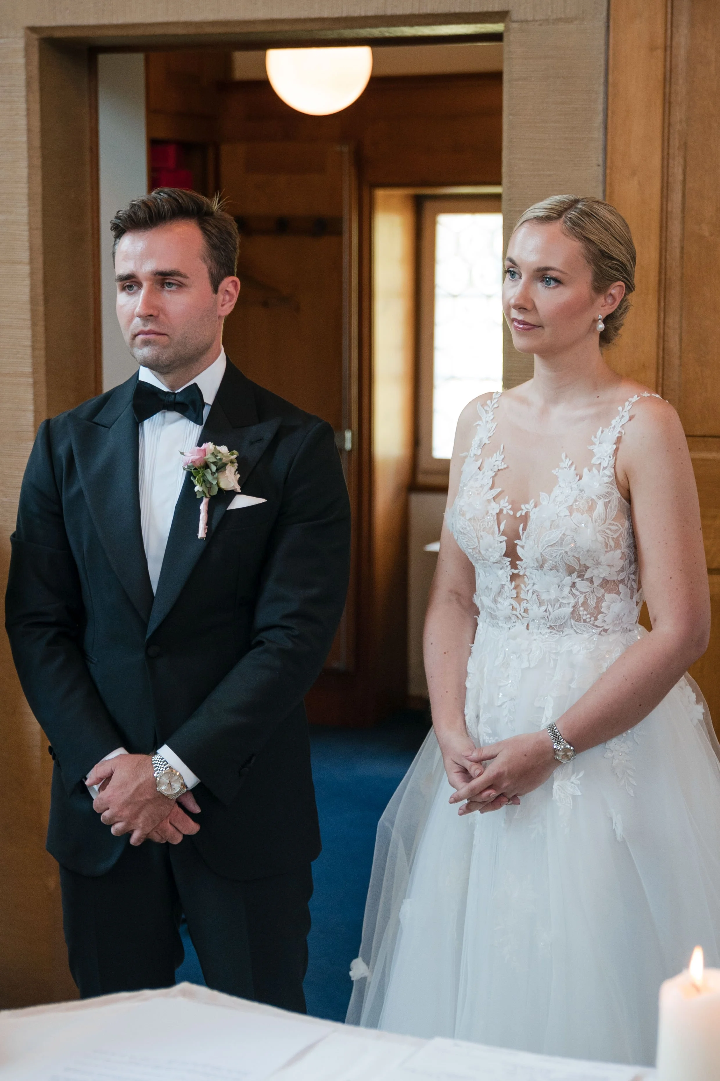 Ein Brautpaar bei einer standesamtlichen Trauung, der Bräutigam trägt einen schwarzen Anzug mit Fliege, die Braut trägt ein weißes Brautkleid mit floraler Spitze und Accessoires. Sie stehen nebeneinander in einem Raum mit Holzdekoration.