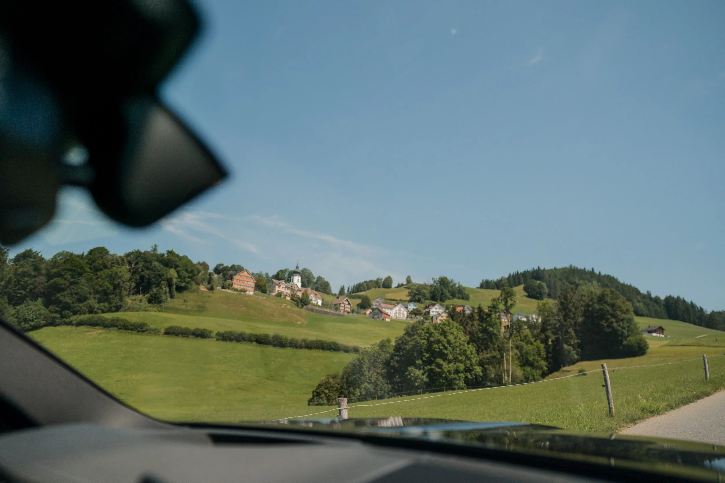 Landschaft mit grünen Hügeln, Bäumen und Häusern, sichtbare Frontscheibe und Spiegel eines Autos