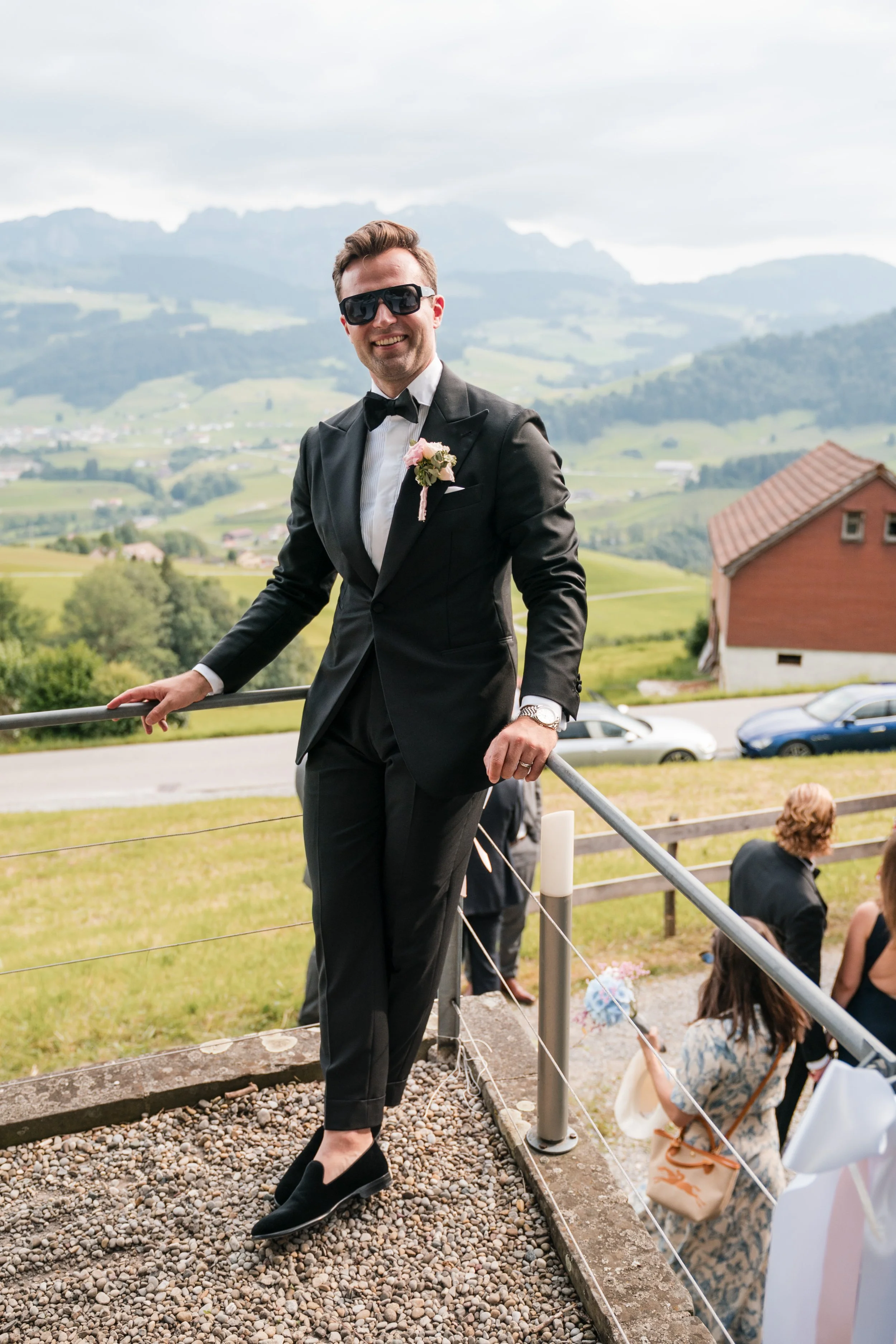 Ein lächelnder Mann in einem schwarzen Anzug, Sonnenbrille und Fliege steht auf einer kleinen Terrasse mit einem ländlichen Hintergrund. Er trägt eine Blumenboutonniere und hat eine Armbanduhr. Im Hintergrund sind grüne Hügel, Häuser und mehrere Pers