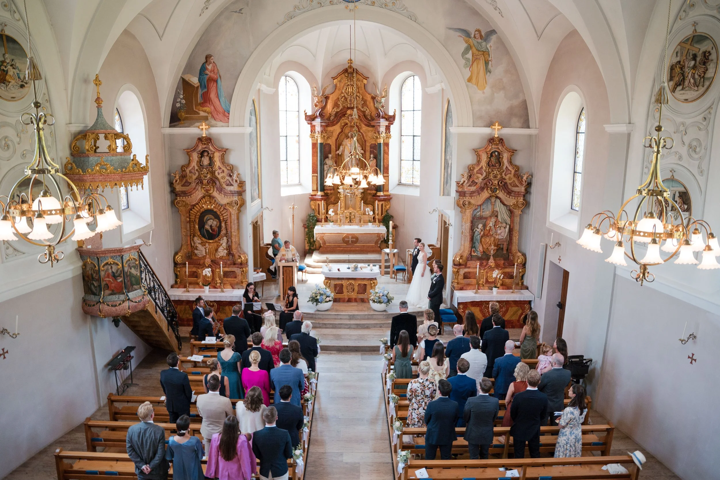 Hochzeitszeremonie in einer Kirche mit vielen Gästen, einem Brautpaar und Geistlichen am Altar, reich verzierte Barockkunst und bunte Fenster im Hintergrund.