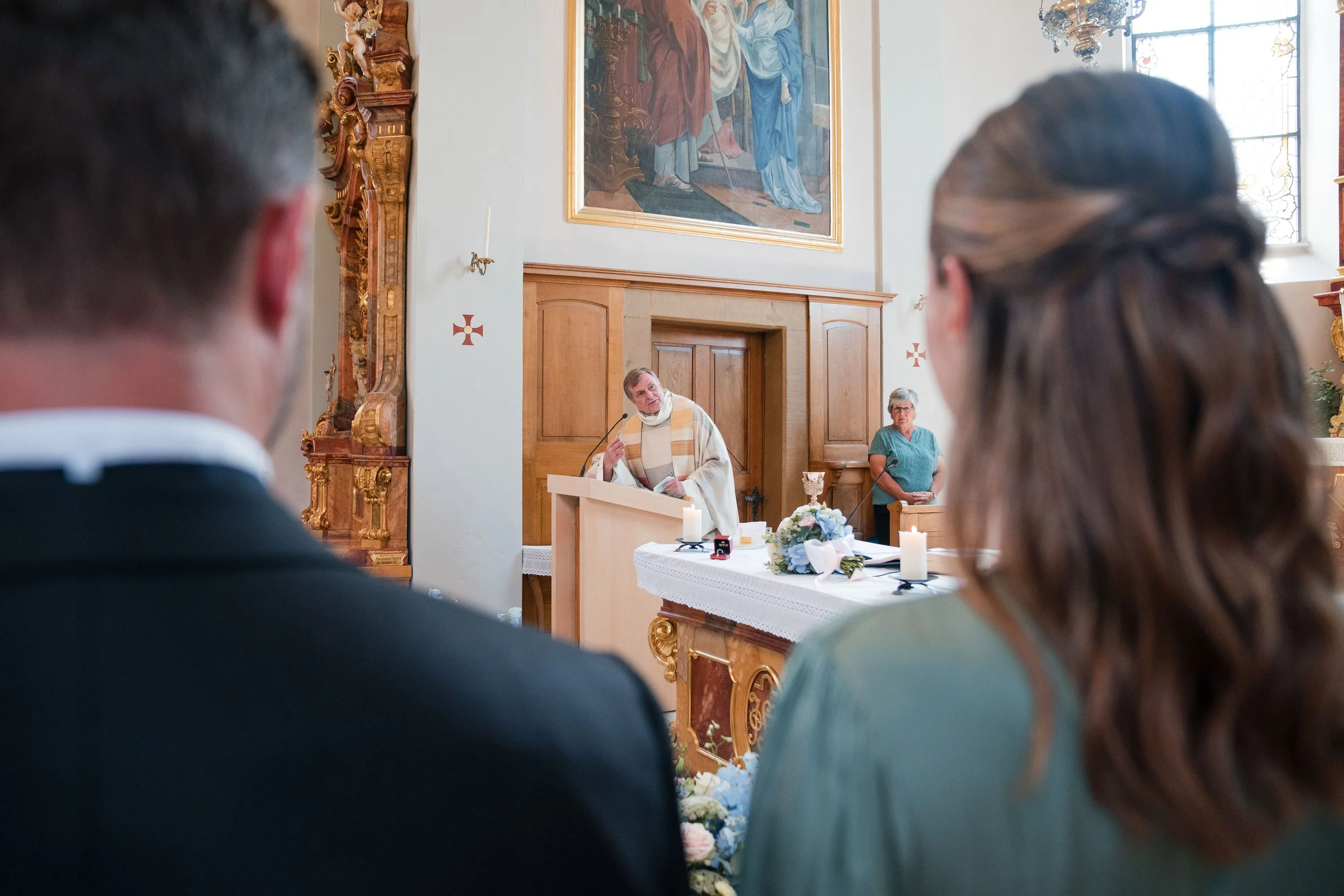 Hochzeitszeremonie in einer Kirche mit einem Priester, einem Brautpaar und einer Frau im Hintergrund.