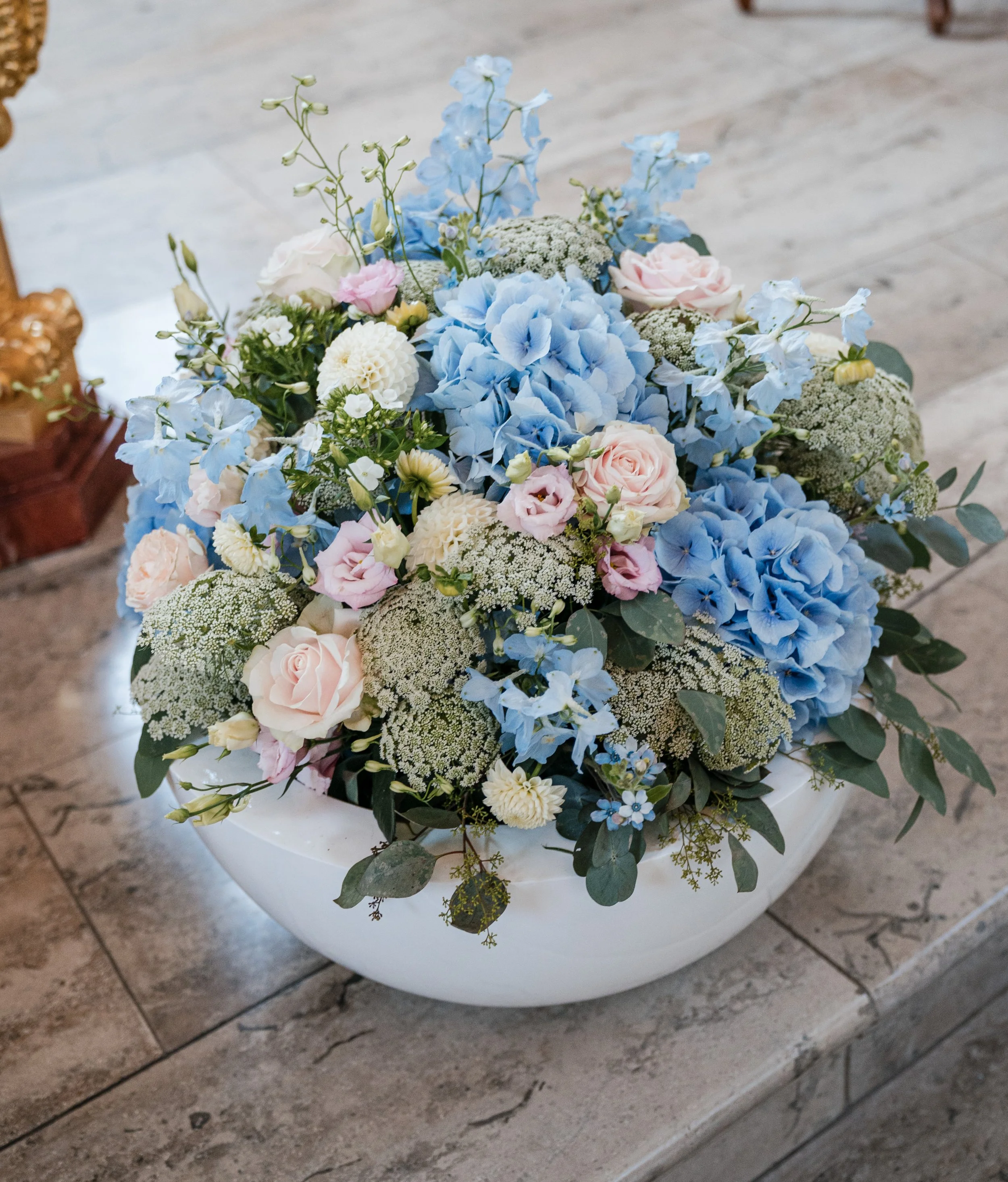 Blumenstrauß mit blauen Hortensien, weißen und rosa Rosen, weißen Dahlien und kleineren Blumen in einer weißen Schale auf einem Steintisch.