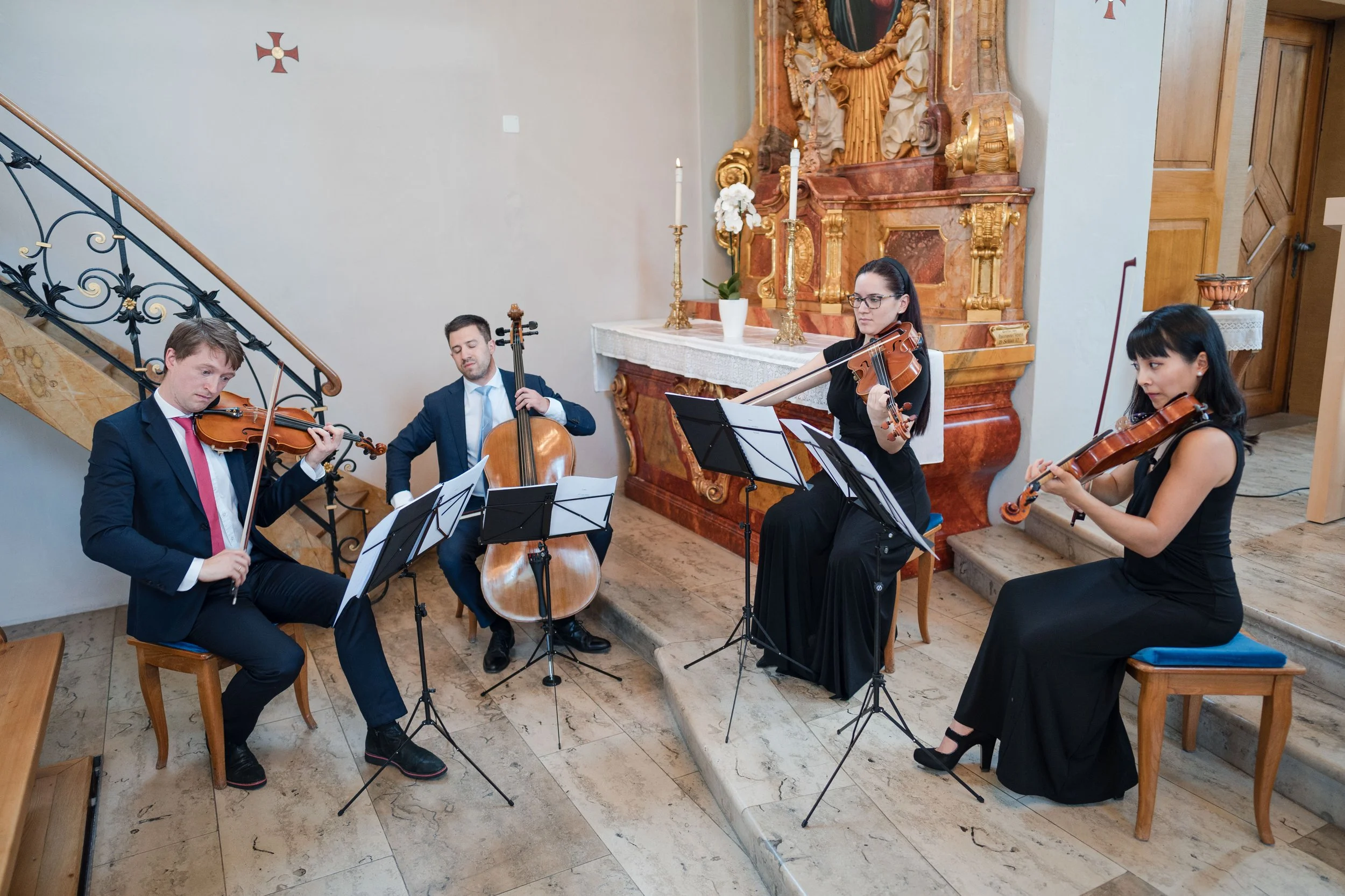 Vier Musiker spielen im Kirchenraum: zwei Violinspielerinnen, ein Cellist und ein Klavierspieler. Sie sind in formeller Kleidung, und im Hintergrund ist eine barocke Altarkopie mit Kerzen und einer Vase mit weißen Blumen zu sehen.