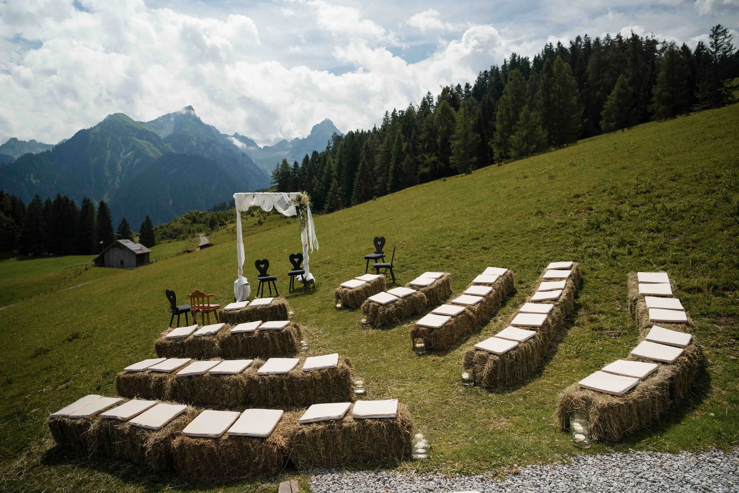 Ein Hochzeitsort im Freien auf einer grasbewachsenen, hügeligen Landschaft mit Bergen im Hintergrund. Es gibt einen weißen Hochzeitsbogen, Sitzgelegenheiten auf Strohballen und Kerzenlichter.