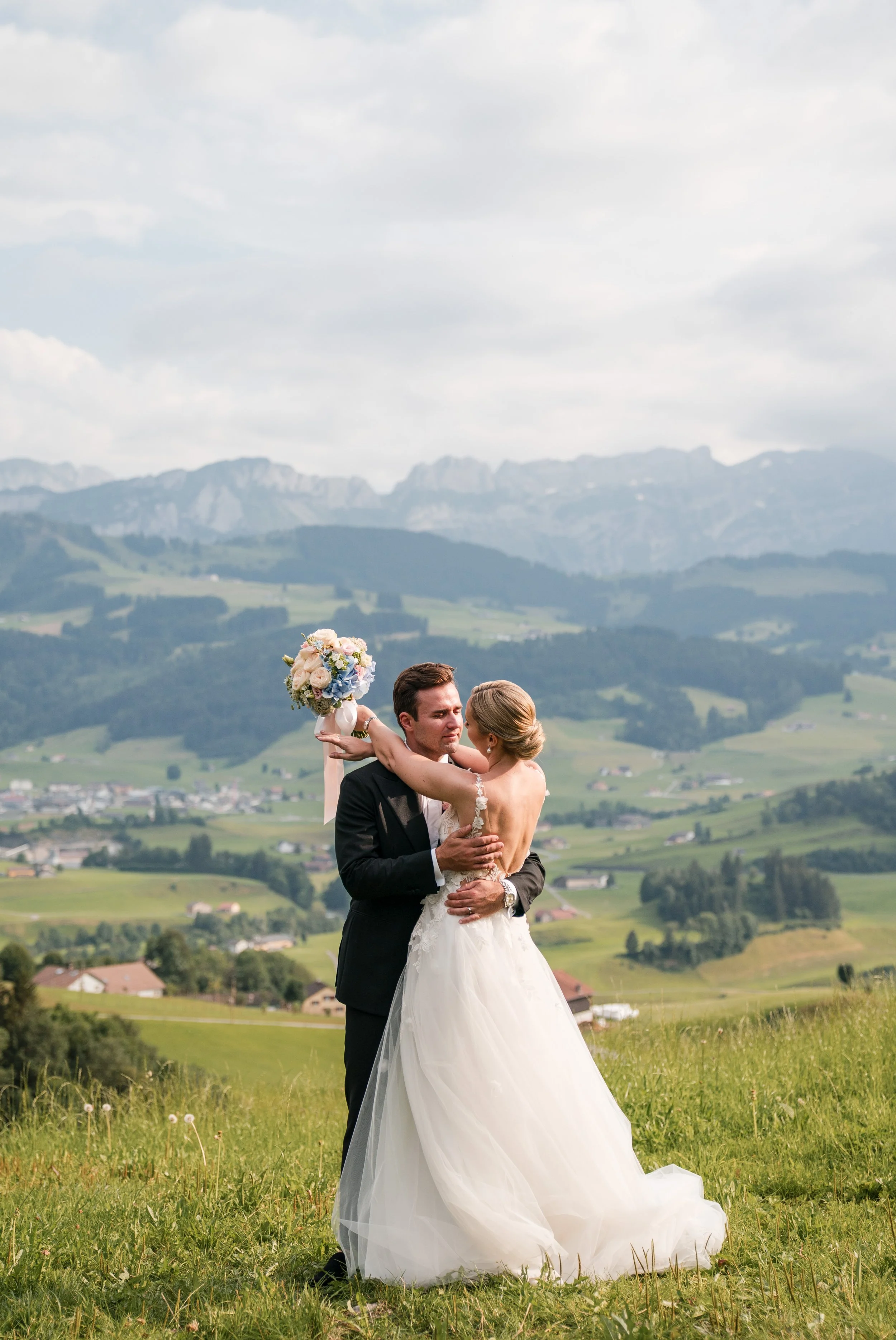 Ein Brautpaar umarmt sich auf einer grünen Wiese mit Bergen im Hintergrund, das Mädchen hält einen Blumenstrauß.